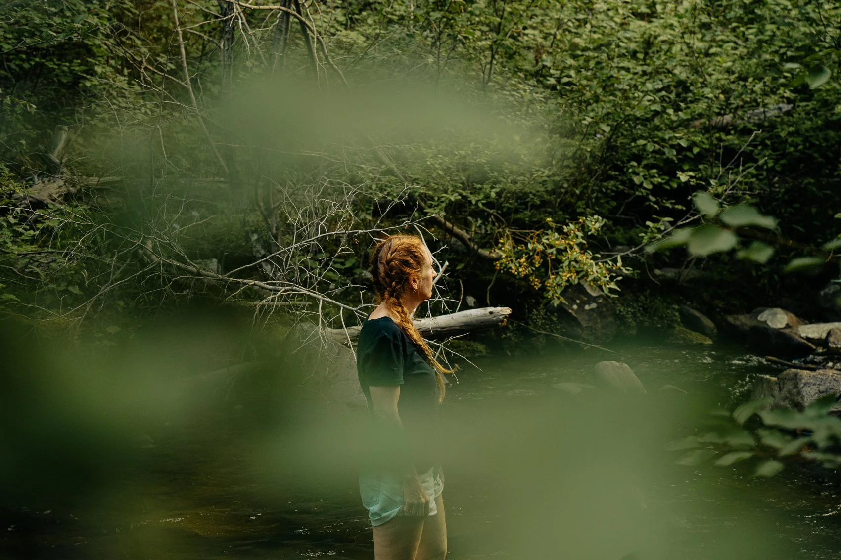 A woman with red hair braided into pigtails standing in a shallow stream surrounded by dense green foliage.