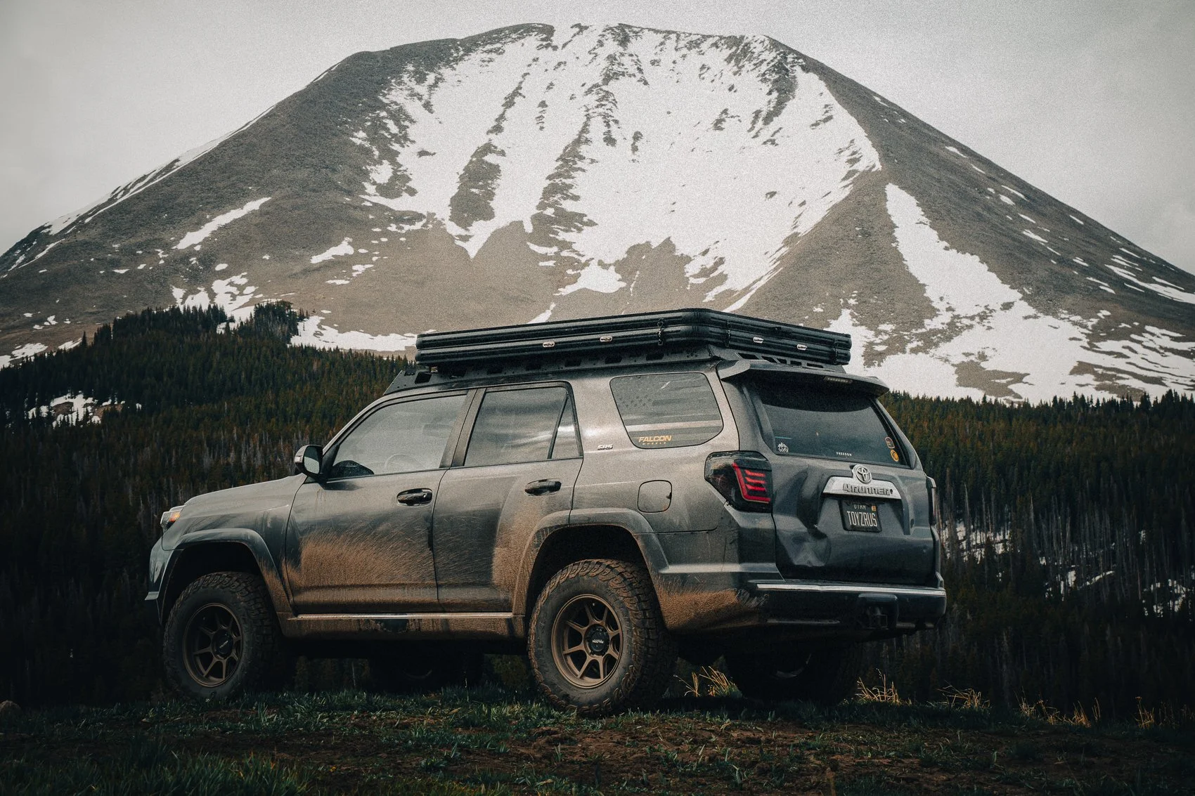 A gray Toyota SUV with off-road tires and a roof rack parked in front of a snow-capped mountain and a forest of evergreen trees.