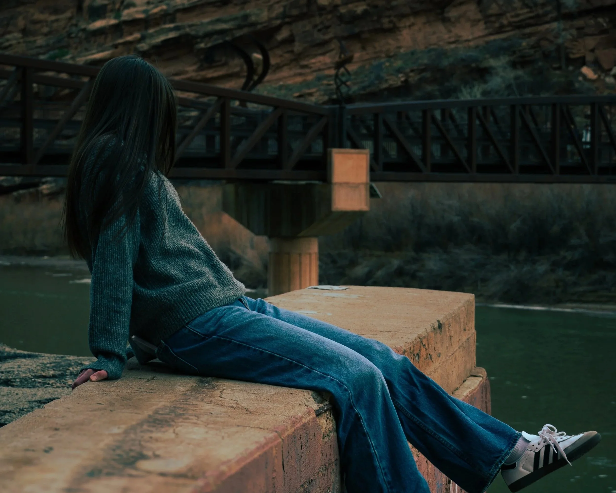 A person with long dark hair sitting on a stone ledge near a river, dressed in a gray sweater, blue jeans, and white sneakers, looking toward the water.
