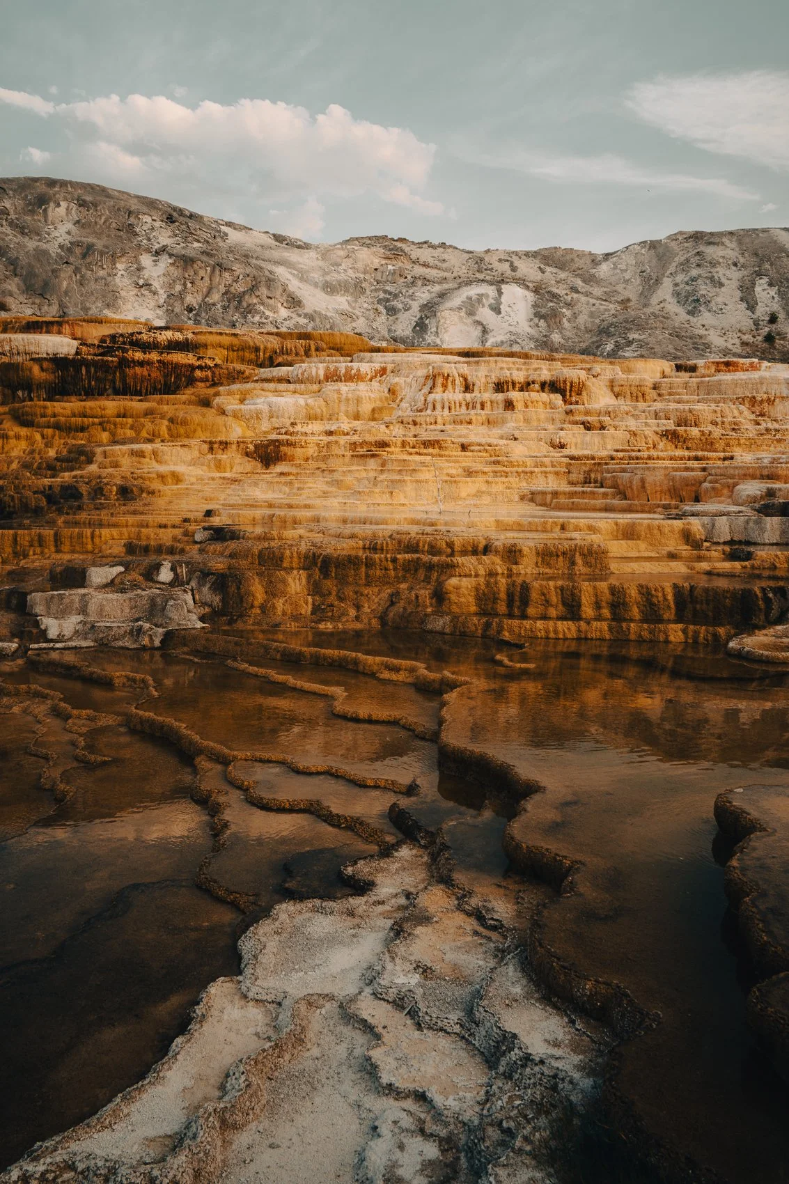 Thermal mineral terraces with water cascading over travertine formations, set against mountains and a partly cloudy sky.