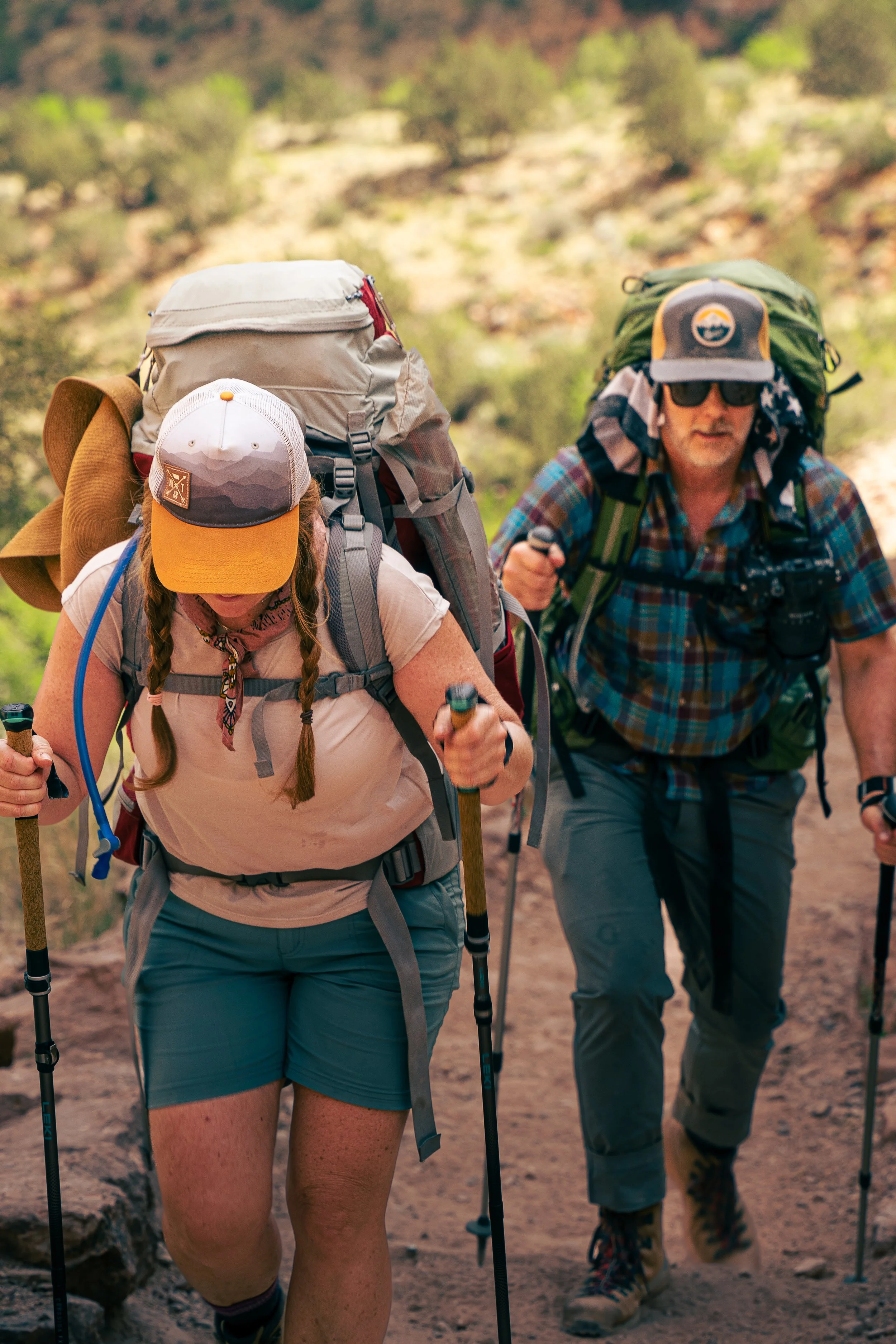 A man and a woman hiking with backpacks and trekking poles in a desert or mountainous area with sparse green vegetation.