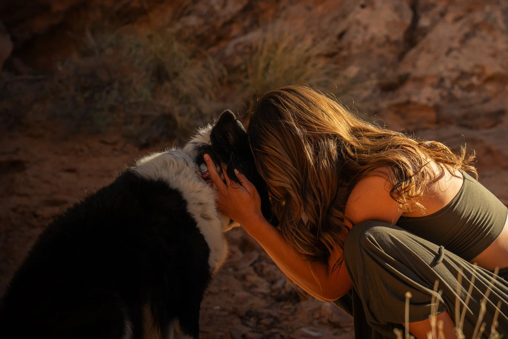 A woman with long wavy hair holding and leaning her forehead against a black and white dog, outdoors in a desert-like area with rocky terrain and warm lighting.