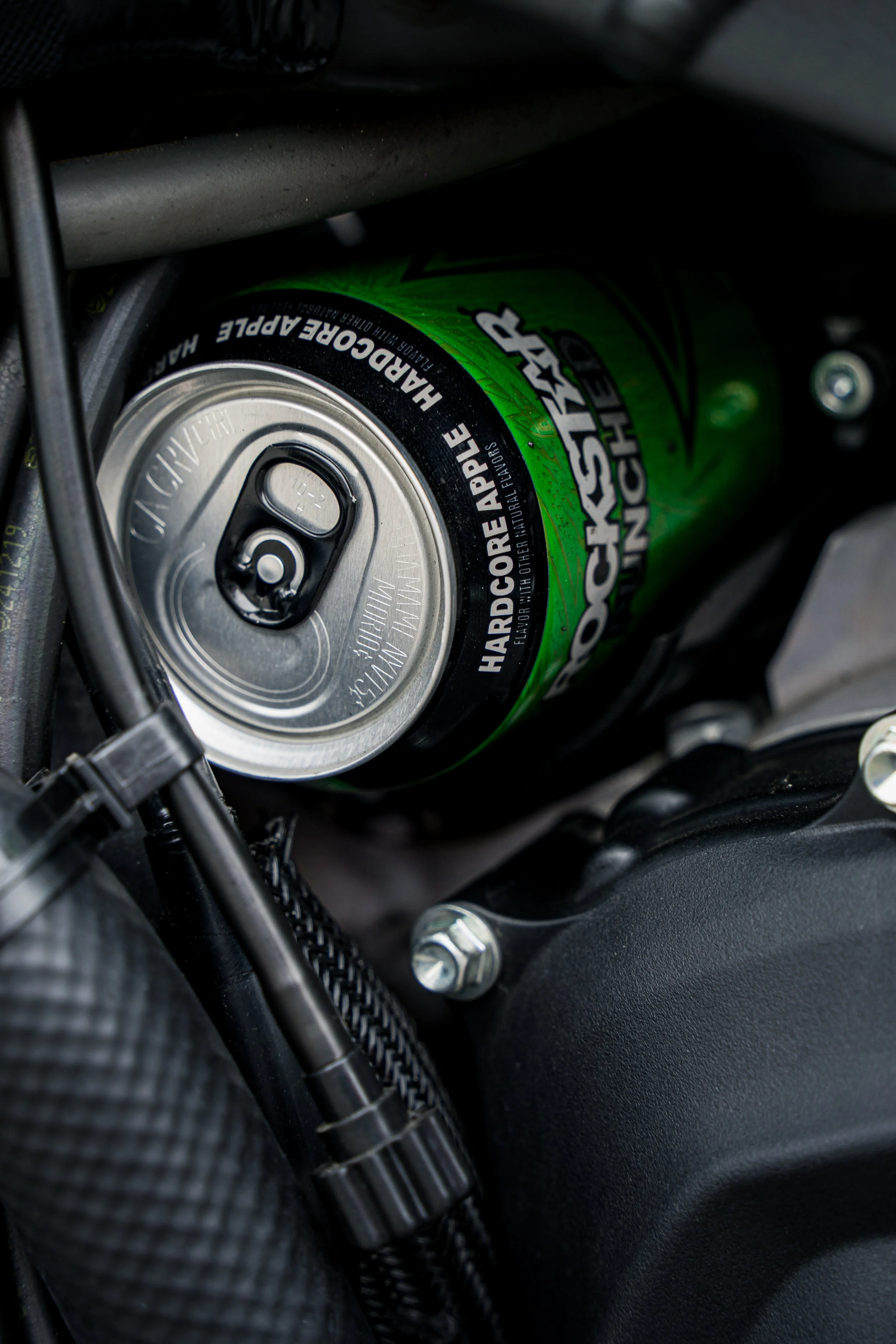 Close-up of a green energy drink can, partially visible, positioned among motorcycle components including a bolt and black textured surface.