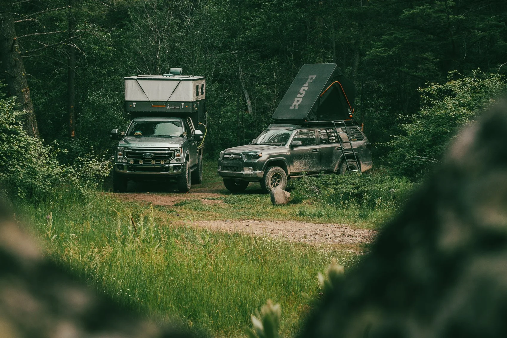 Two off-road vehicles parked in a lush forest clearing, with camping equipment on top, including rooftop tents and solar panels.