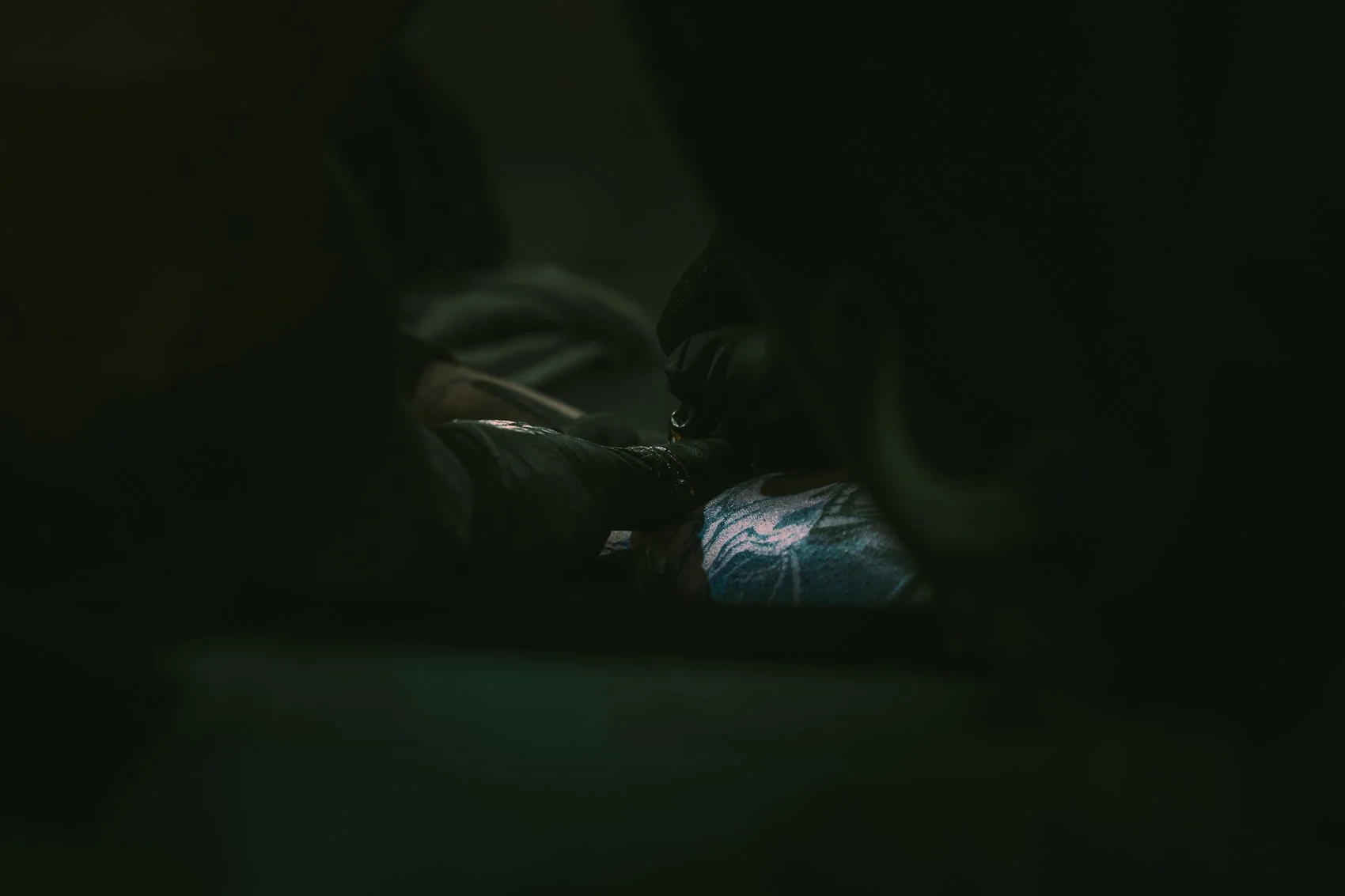 Dark photo showing a person wearing black gloves and shoes, surrounded by darkness with a faintly visible tattoo gun.
