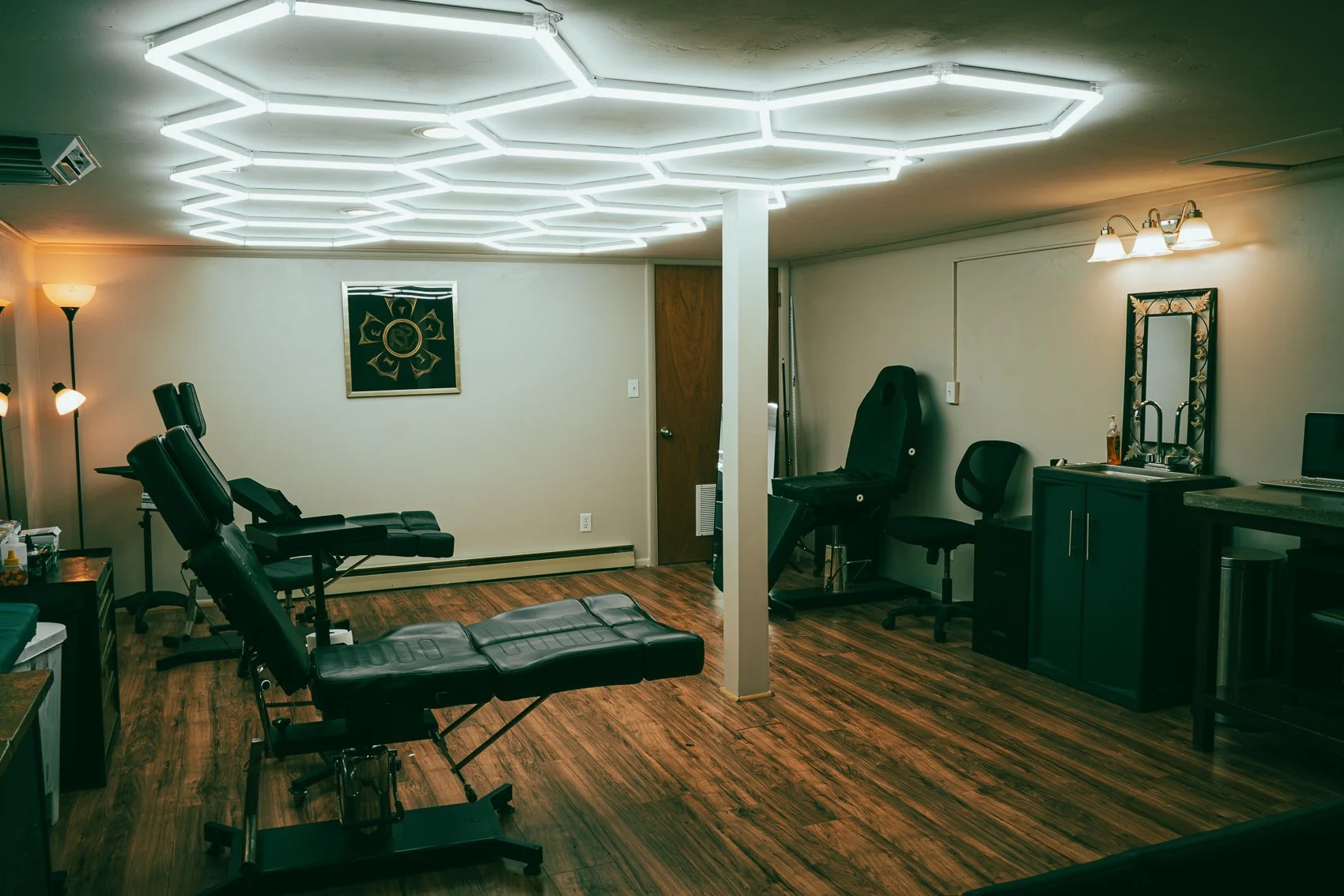 Room with chiropractic or massage equipment, including adjustable massage tables, a chair, a cabinet with a mirror, and lighting fixtures, on wood flooring.