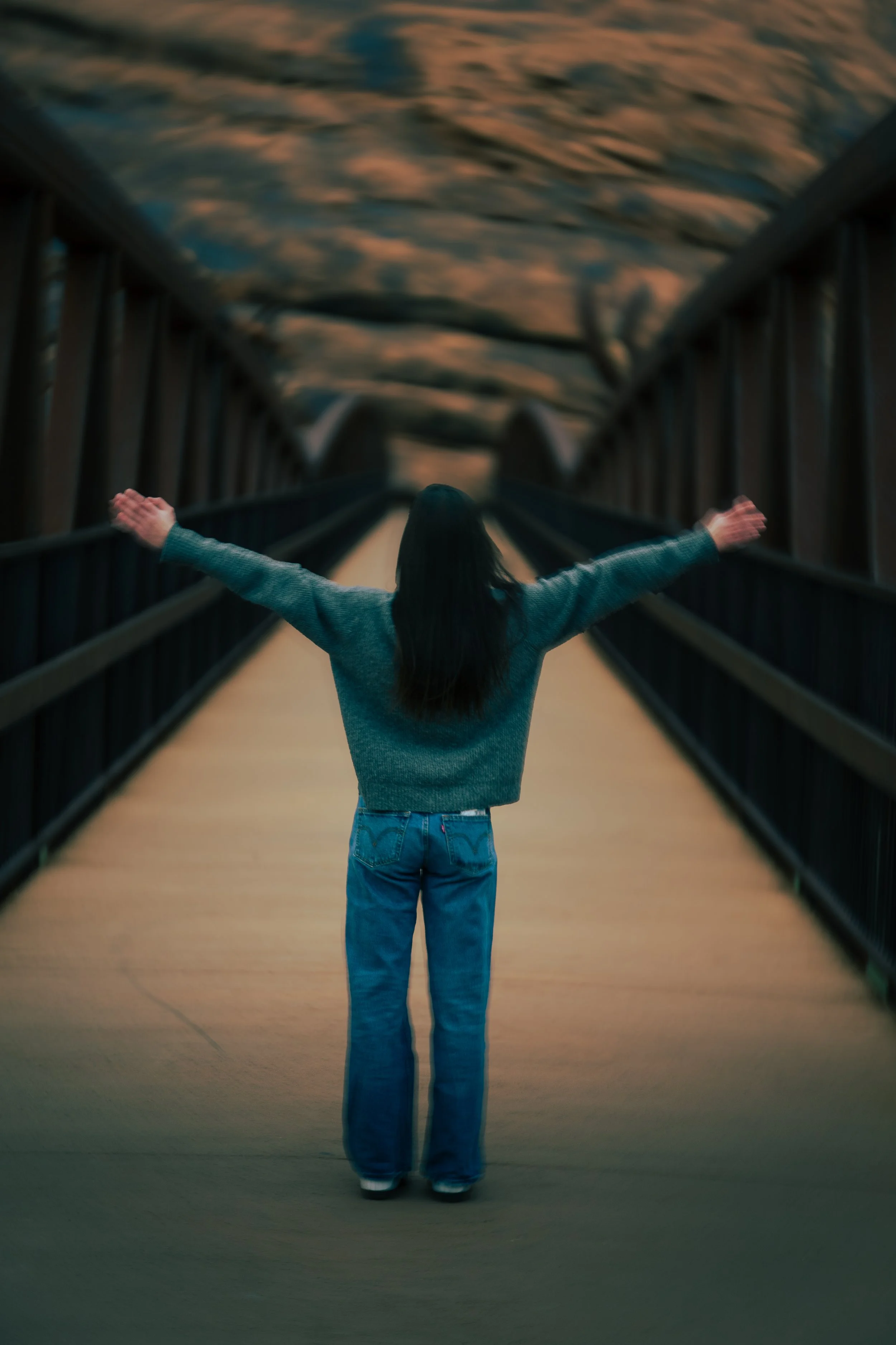 A person with long dark hair wearing a gray sweater and blue jeans standing on a bridge with arms outstretched, facing away, and looking at a cloudy sunset sky.