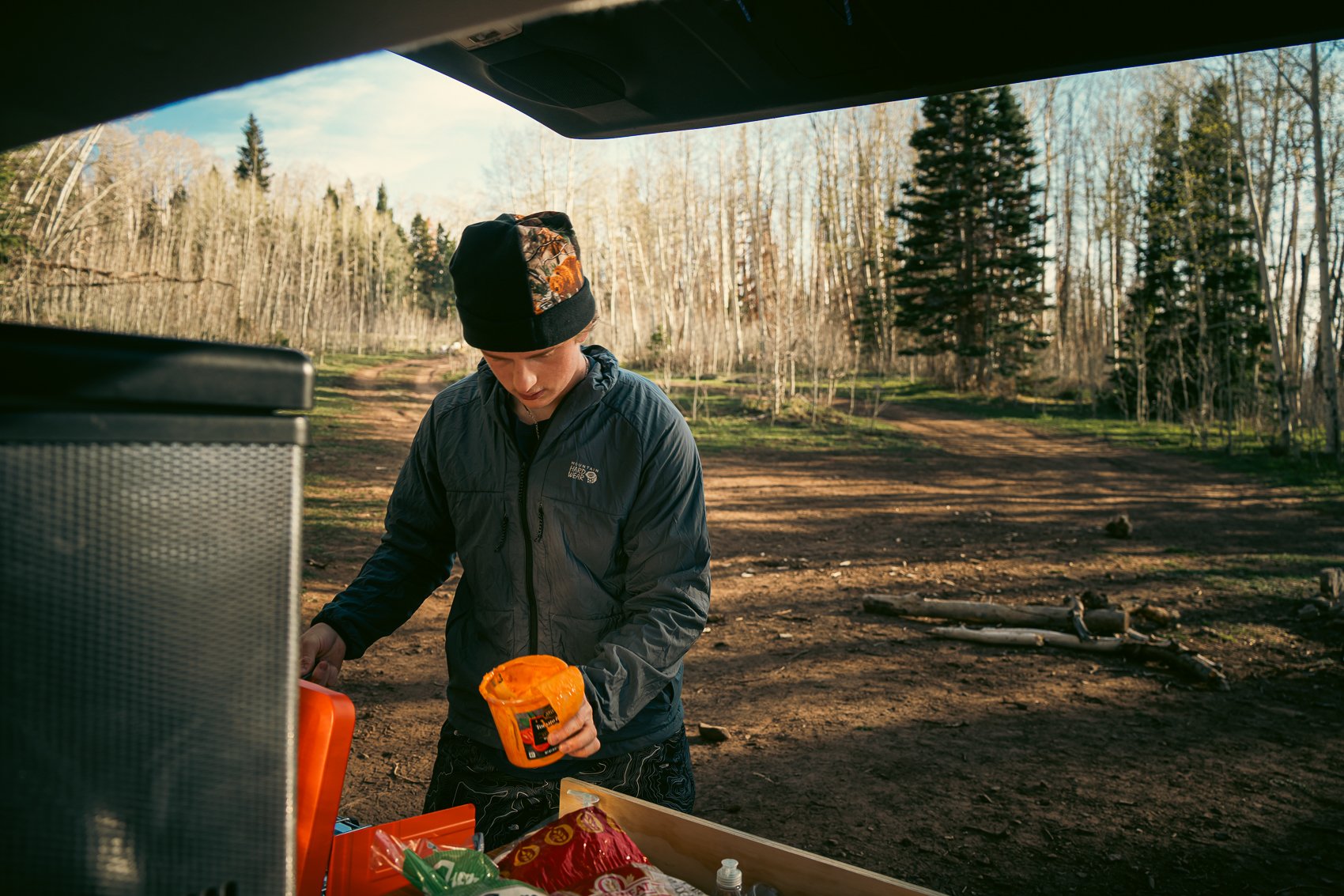 A young person in outdoor gear standing at an organized outdoor cooking or camping station in a forested area, with trees and a dirt trail in the background, preparing snacks or food.