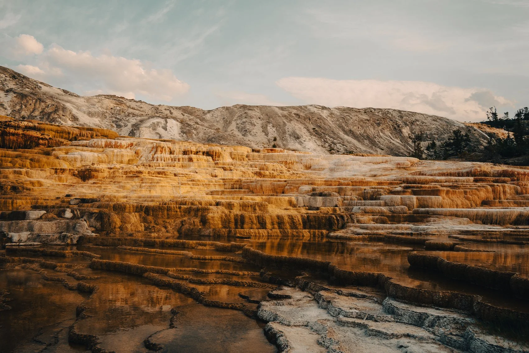 Terraced hot springs with mineral deposits in shades of orange, white, and brown, set against rocky, mountainous terrain under a partly cloudy sky.