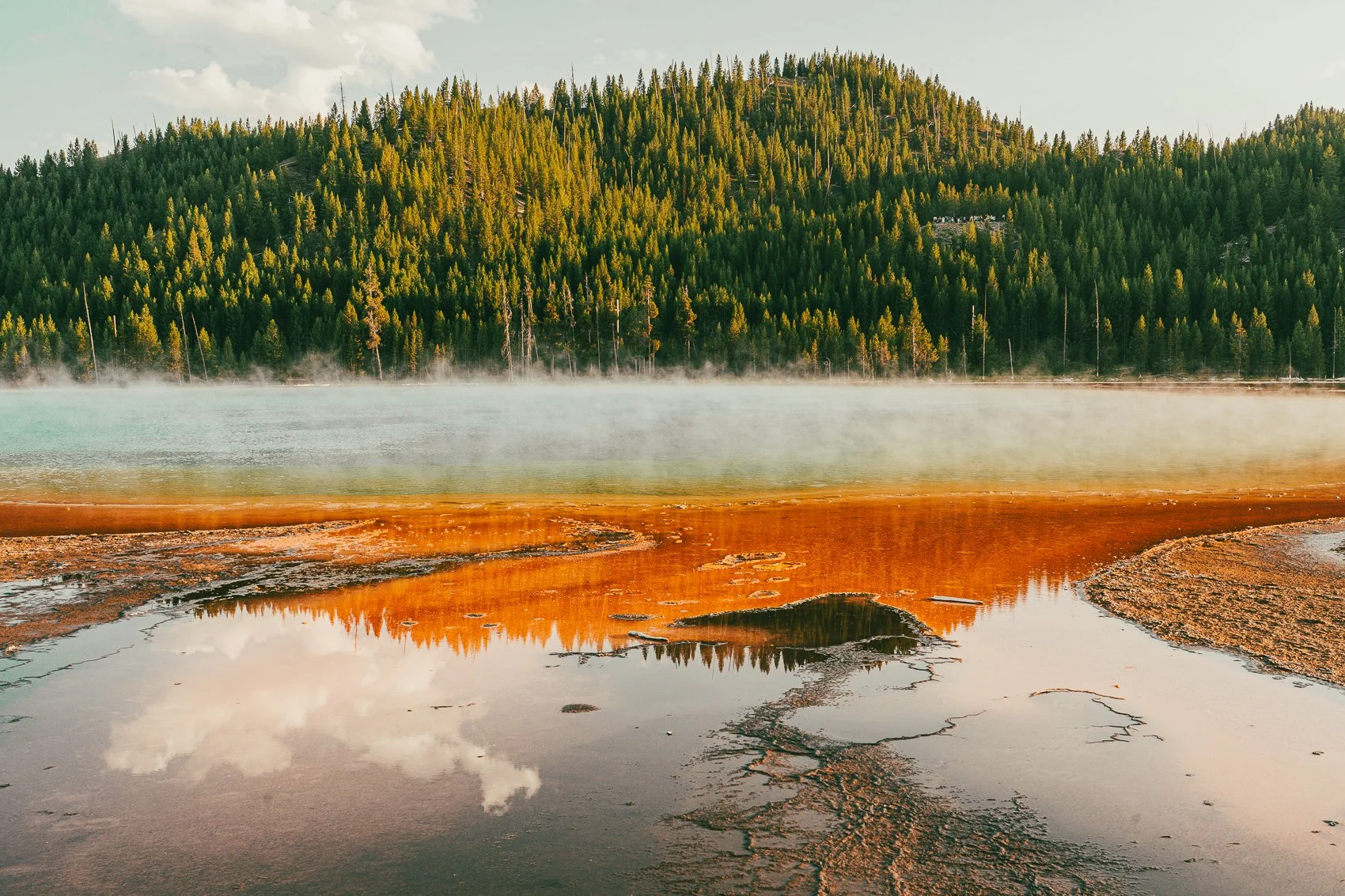 A hot spring with vibrant orange, green, and black mineral deposits, steam rising over the water, and a forested mountain in the background under a partly cloudy sky.