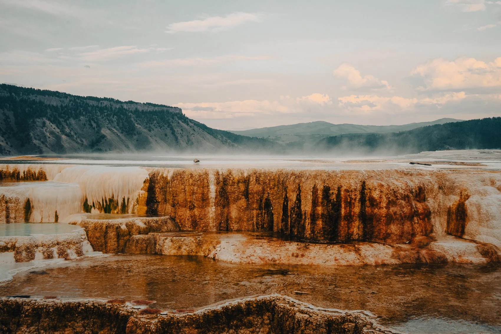 Colorful hot springs with mineral deposits in a geothermal area, surrounded by green mountains under a partly cloudy sky.