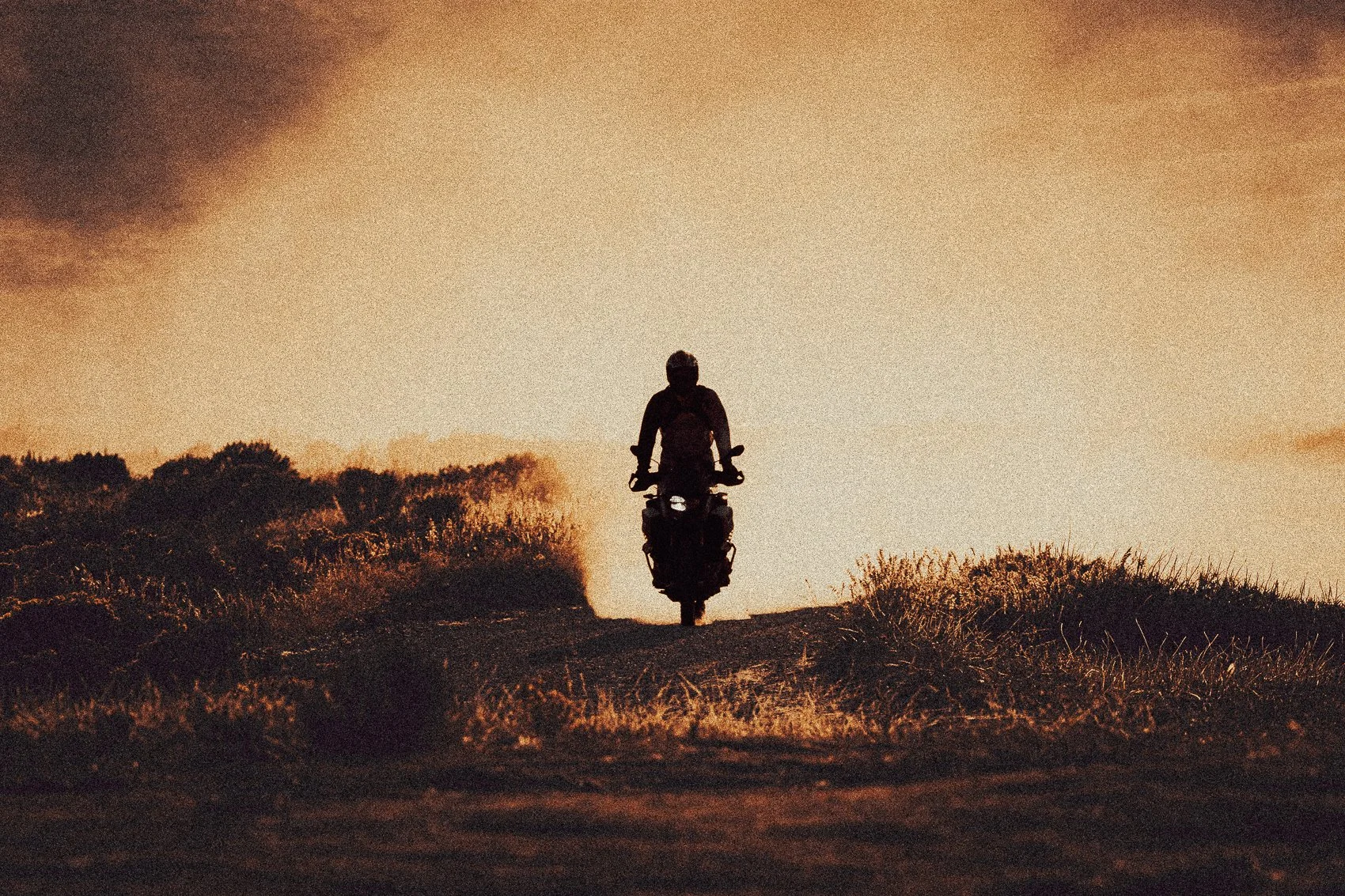 A person riding a motorcycle on a dirt path during sunset, silhouetted against a colorful sky with clouds, surrounded by tall grass and trees.