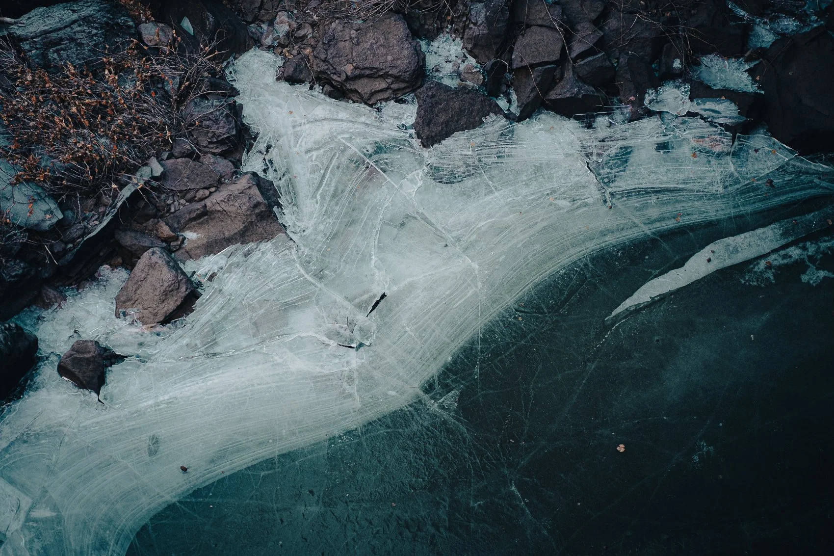 Close-up of a frozen surface with cracked, translucent ice and dark rocks underneath, along with some dried plants in the background.