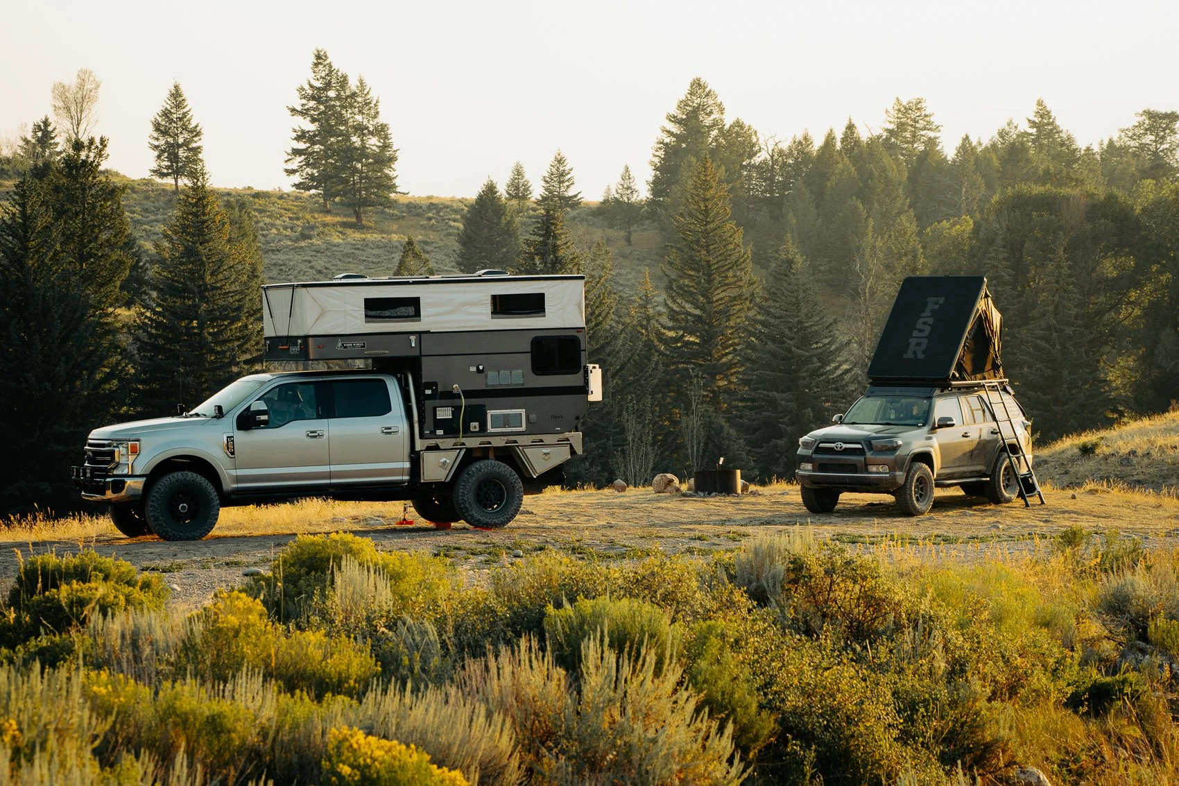 Two off-road vehicles parked in a grassy field surrounded by trees, one with a rooftop tent and the other with an foor wheel campers camper.
