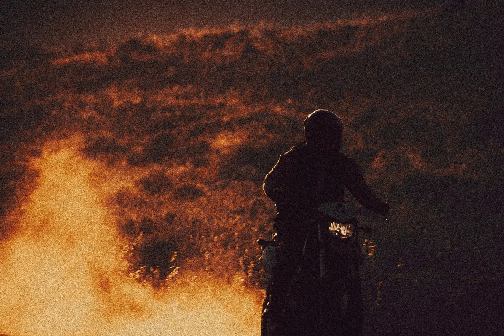 Person riding a motorcycle at night with a fiery background and dark sky.