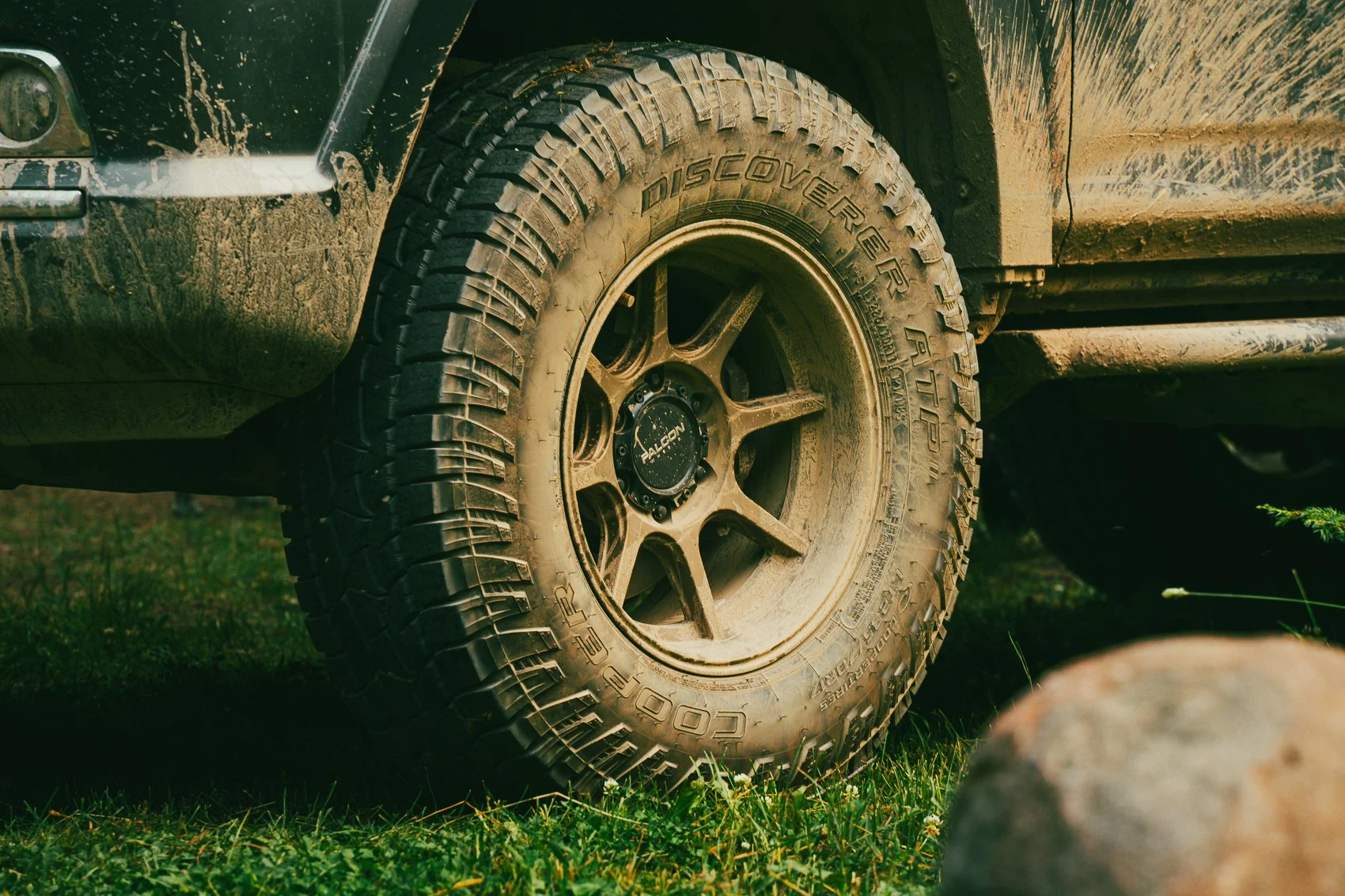 Close-up of a dirty off-road vehicle tire with 'Discoverer' written on the sidewall, mounted on a metallic wheel with a black center cap. The vehicle has mud splatters on the body near the tire, and the ground is grassy with rocks nearby.