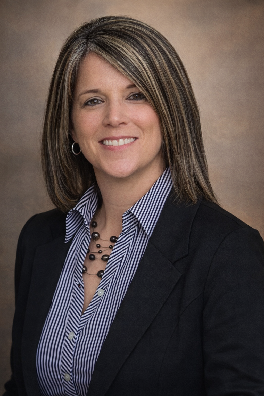 A professional woman with shoulder-length brown hair with blonde highlights, smiling, wearing a black blazer over a striped button-up shirt, and a beaded necklace and hoop earrings.