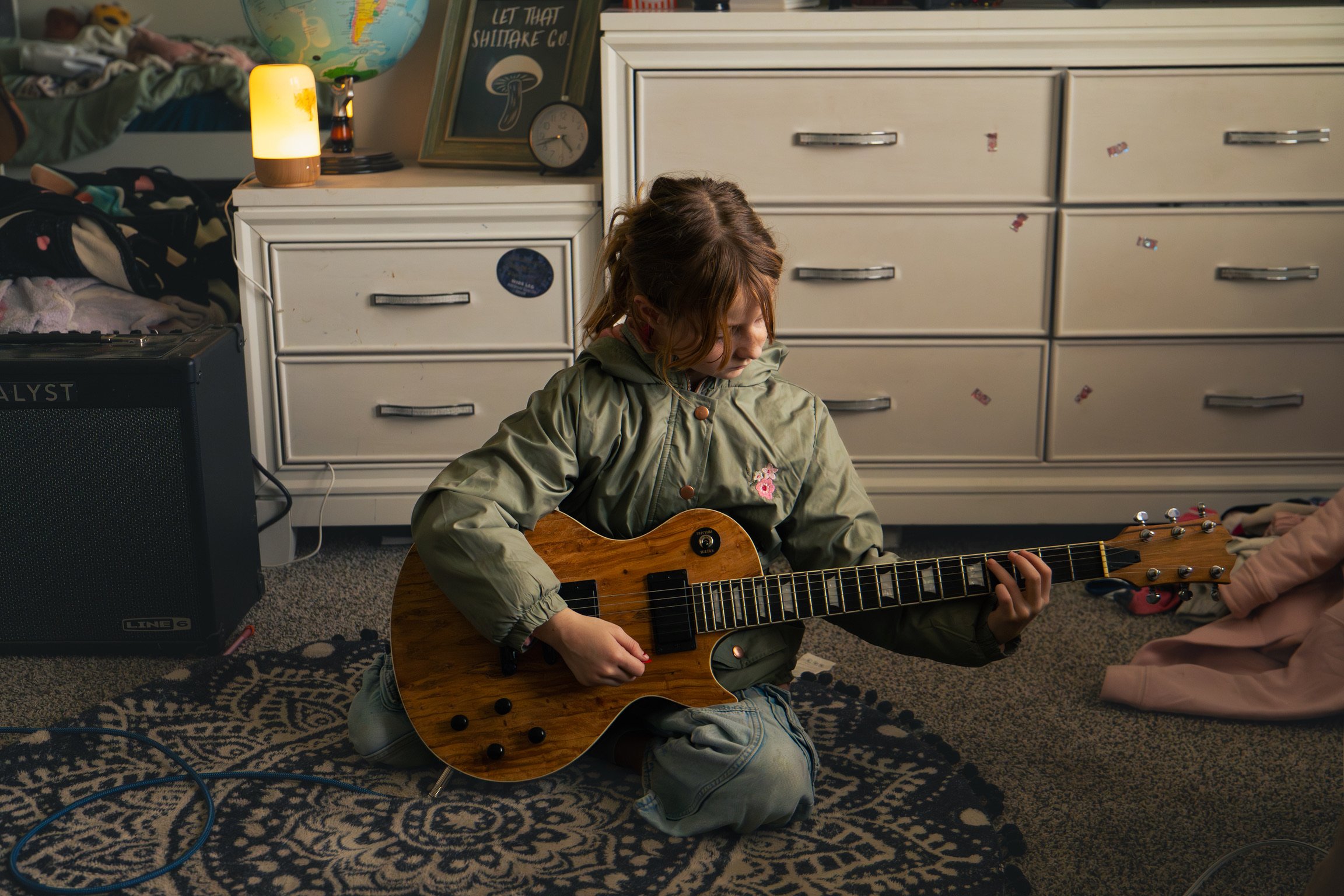 A young girl with red hair kneels on a patterned rug, playing an acoustic guitar indoors. The room has white furniture, including a dresser with stickers, a lamp, a globe, and a framed mushroom poster. Clothes are scattered on a nearby bed.