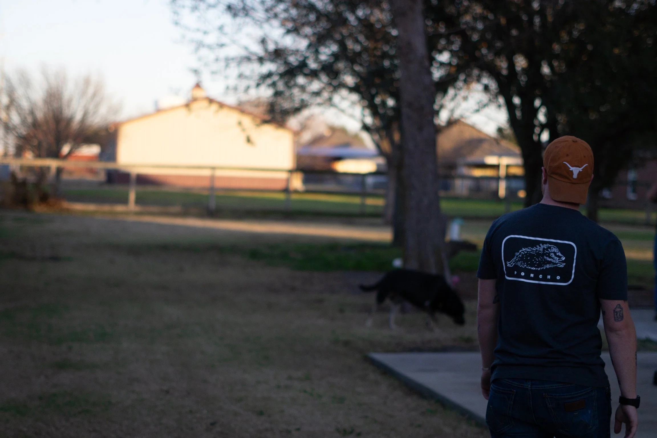 A person wearing a Texas Longhorns hat and a black T-shirt with a graphic design on the back, walking outdoors in a park area with trees, a fence, and houses in the background during evening or late afternoon.