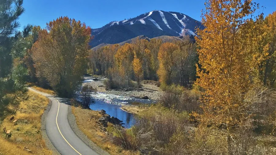 A winding two-lane road running alongside a river with trees showing fall colors, and a snow-capped mountain in the background under a clear blue sky.