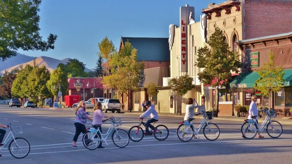 People riding bikes and walking on a city street with shops and trees, with mountains in the background.