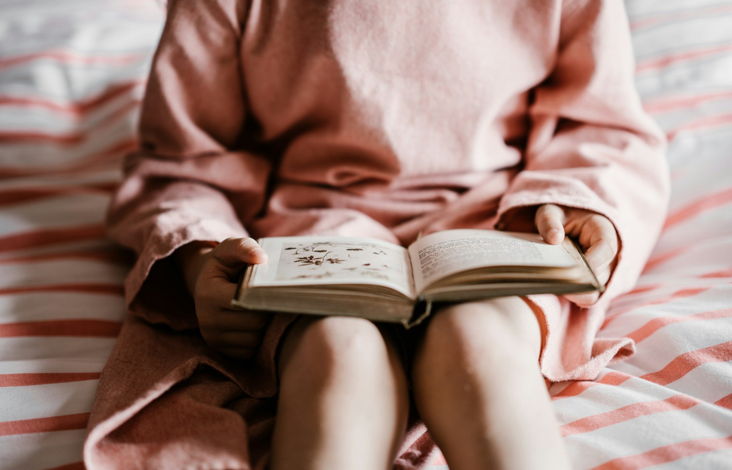 Child in pink pajamas sitting on bed, reading a book with botanical illustrations.