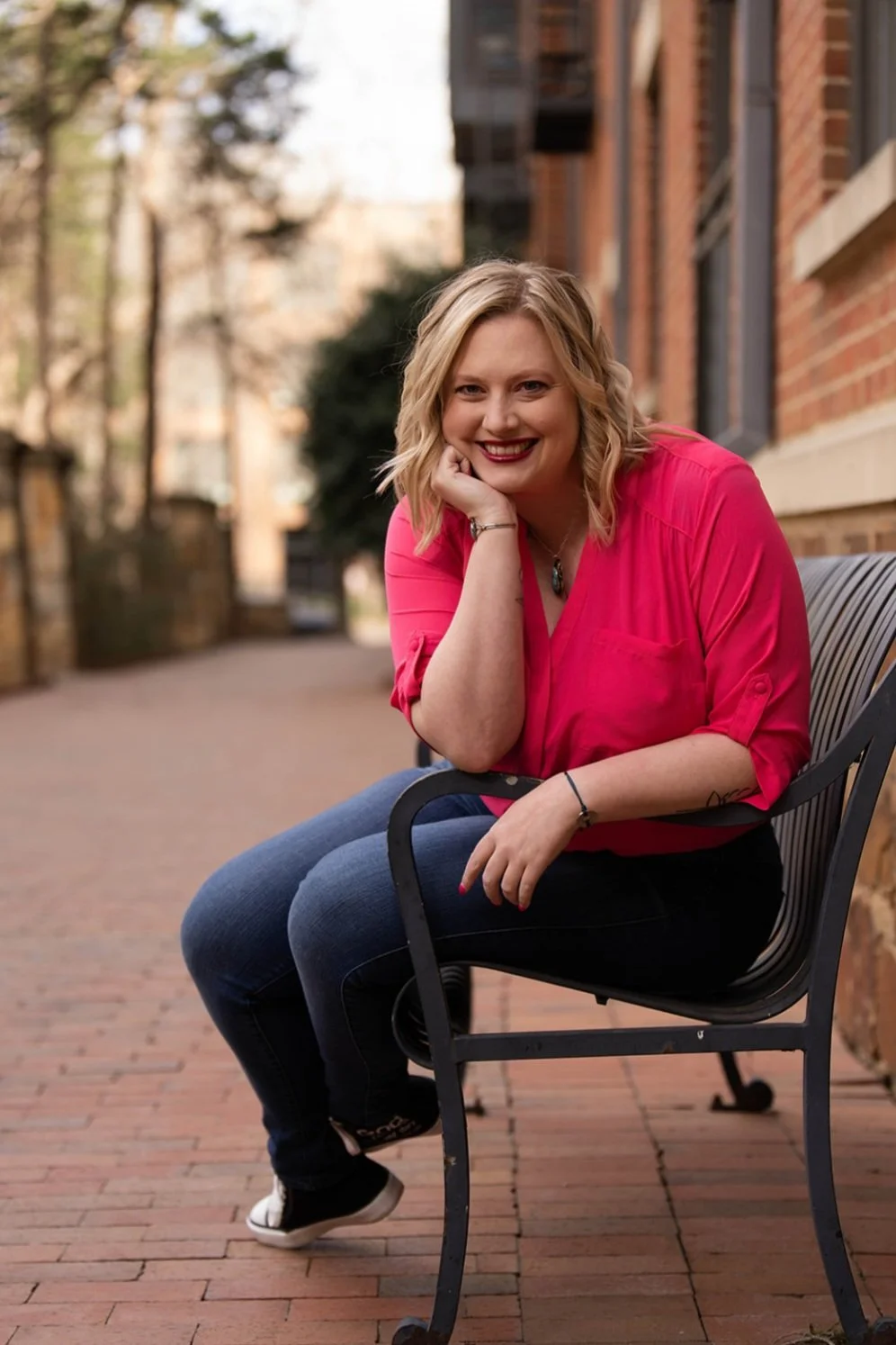 A woman with blonde hair wearing a bright pink blouse and dark jeans sitting on a black metal bench outside on a brick-paved area, smiling at the camera with her hand resting on her chin.