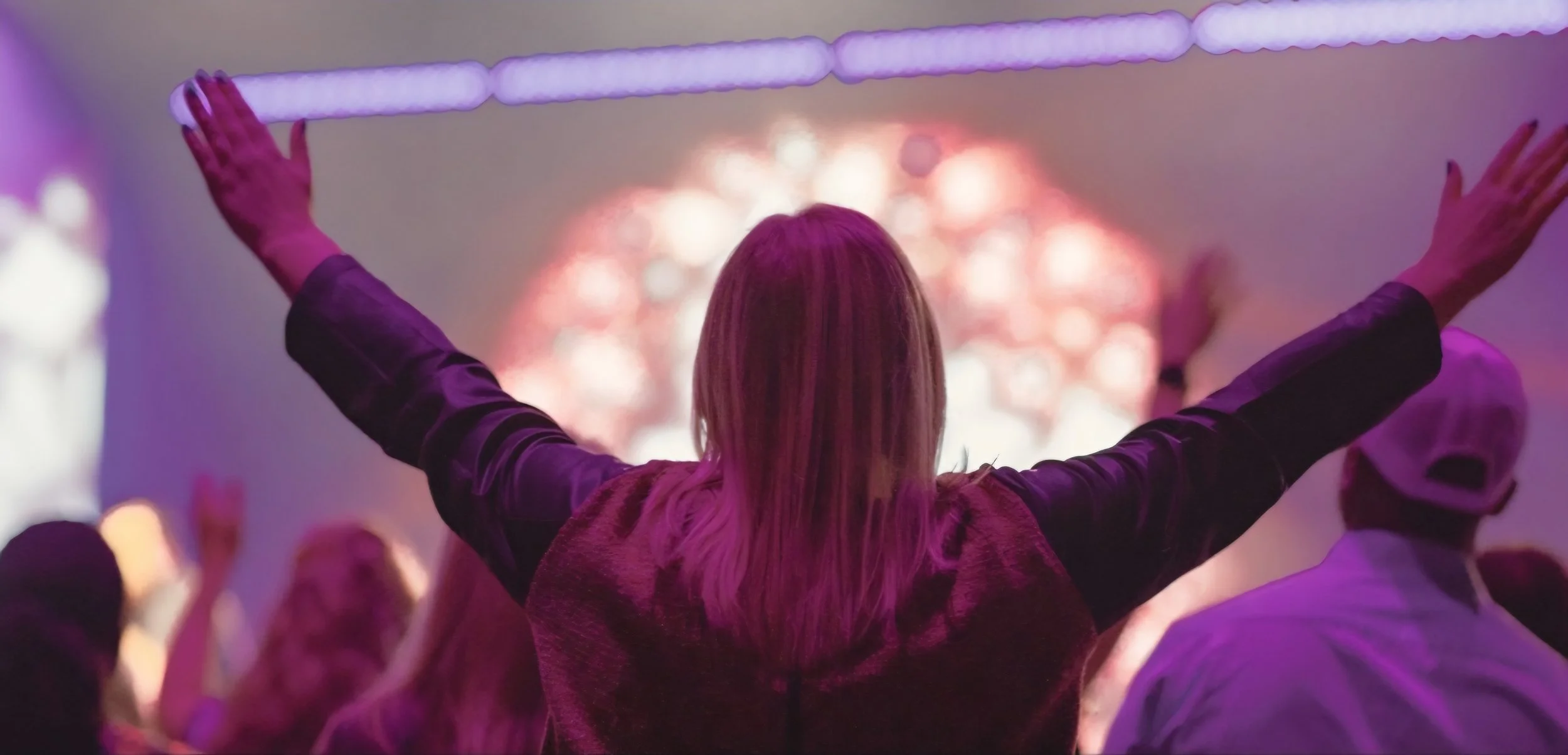 A woman with long blonde hair standing with arms raised holding a light stick at a concert or event, viewed from behind, with colorful lights and a crowd in the background.