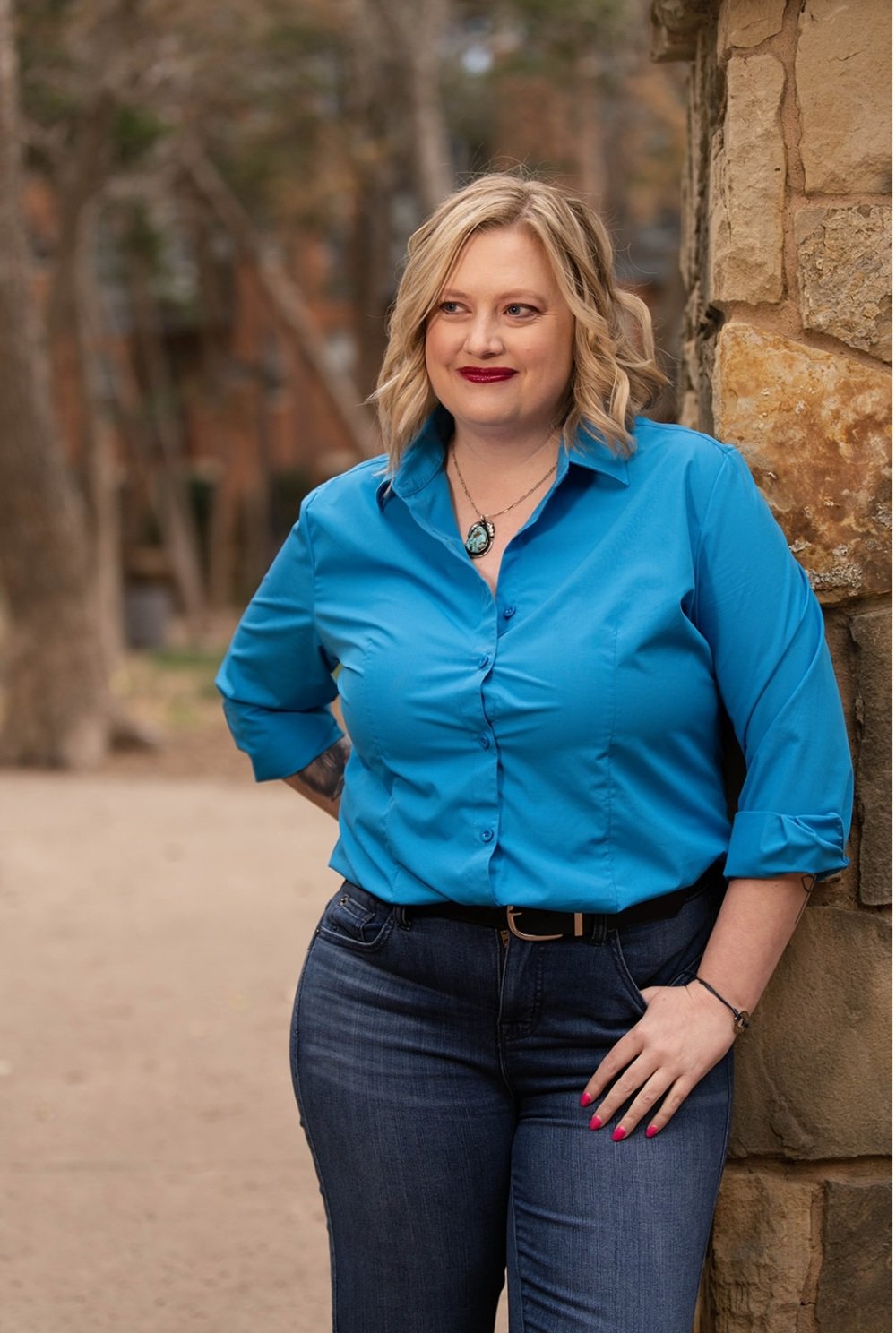 A woman with blonde, wavy hair standing outdoors near a stone wall, wearing a bright blue shirt with rolled-up sleeves and dark jeans, smiling softly and looking to her left.