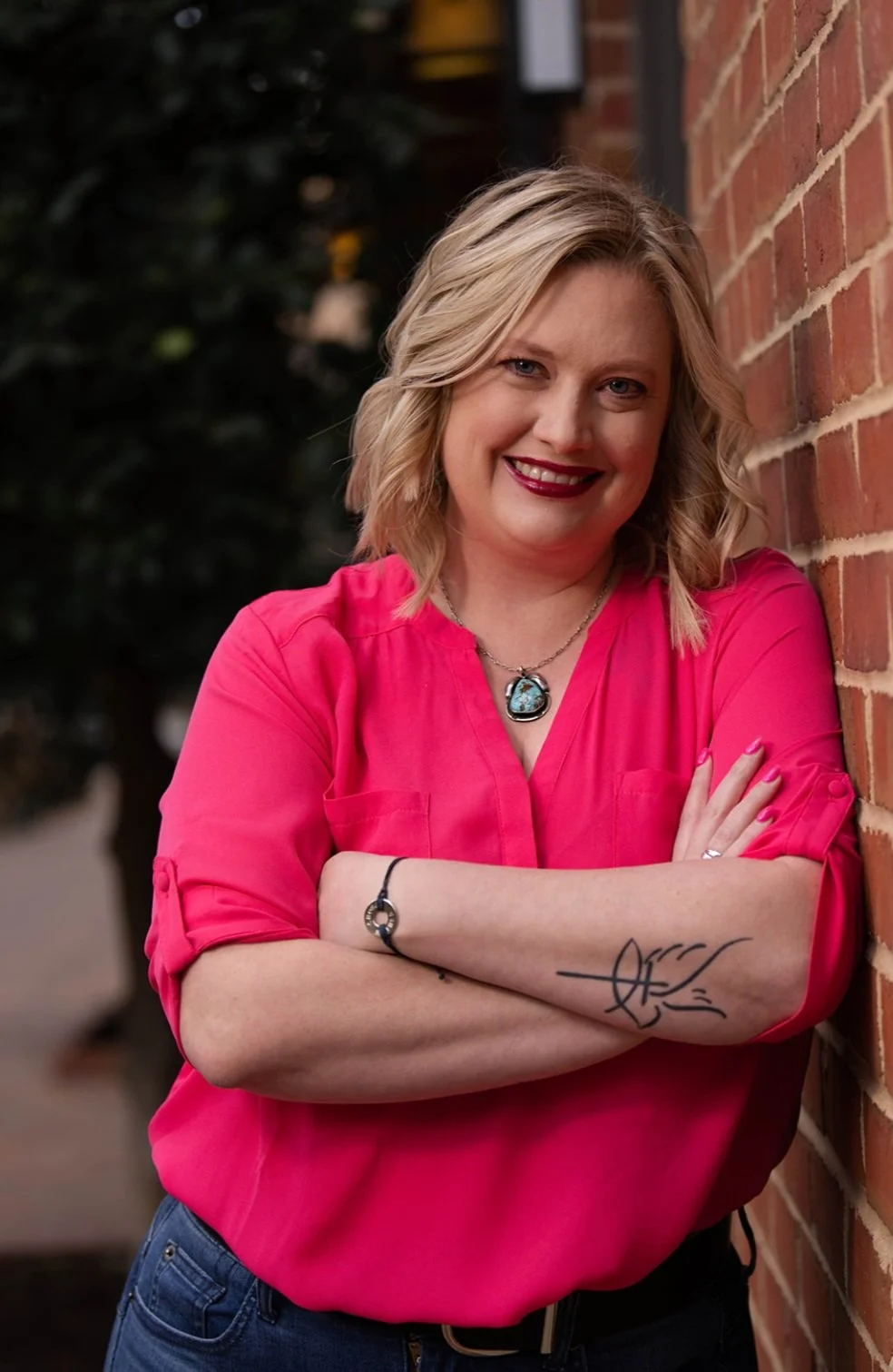 A smiling woman with blonde wavy hair, wearing a bright pink blouse, standing against a brick wall with her arms crossed, showing a tattoo on her forearm and wearing jewelry.