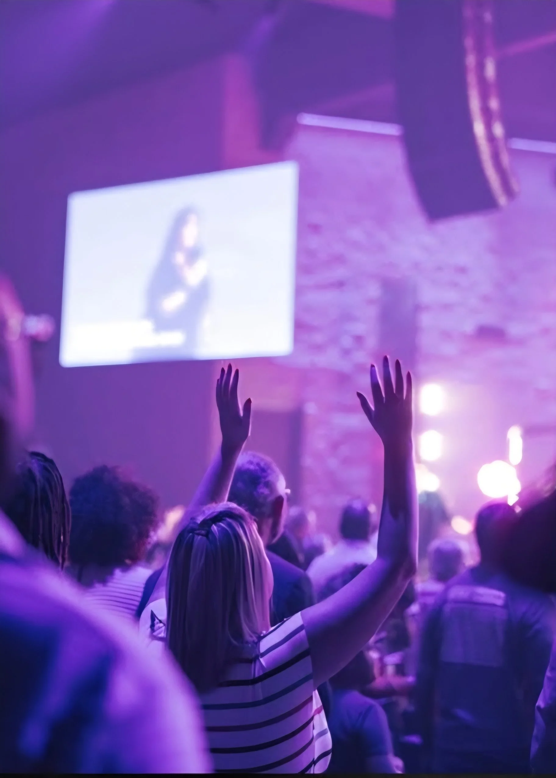 Crowd of people with hands raised in a concert or live event, with purple lighting, large screen displaying a performer on stage, and speakers in the background.