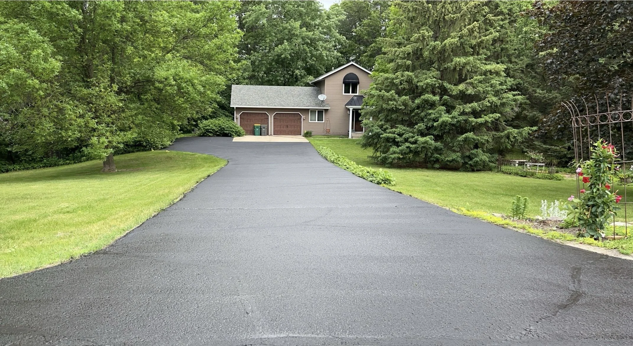 A sealed driveway leading to a two-story house surrounded by green trees and a well-maintained lawn.