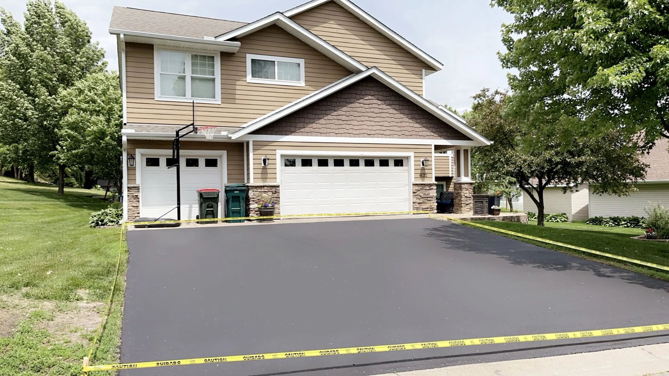 Freshly sealed driveway with caution tape around it in front of a beige, two-story house with white garage doors, green trash cans, and a basketball hoop, surrounded by green trees and lawn.