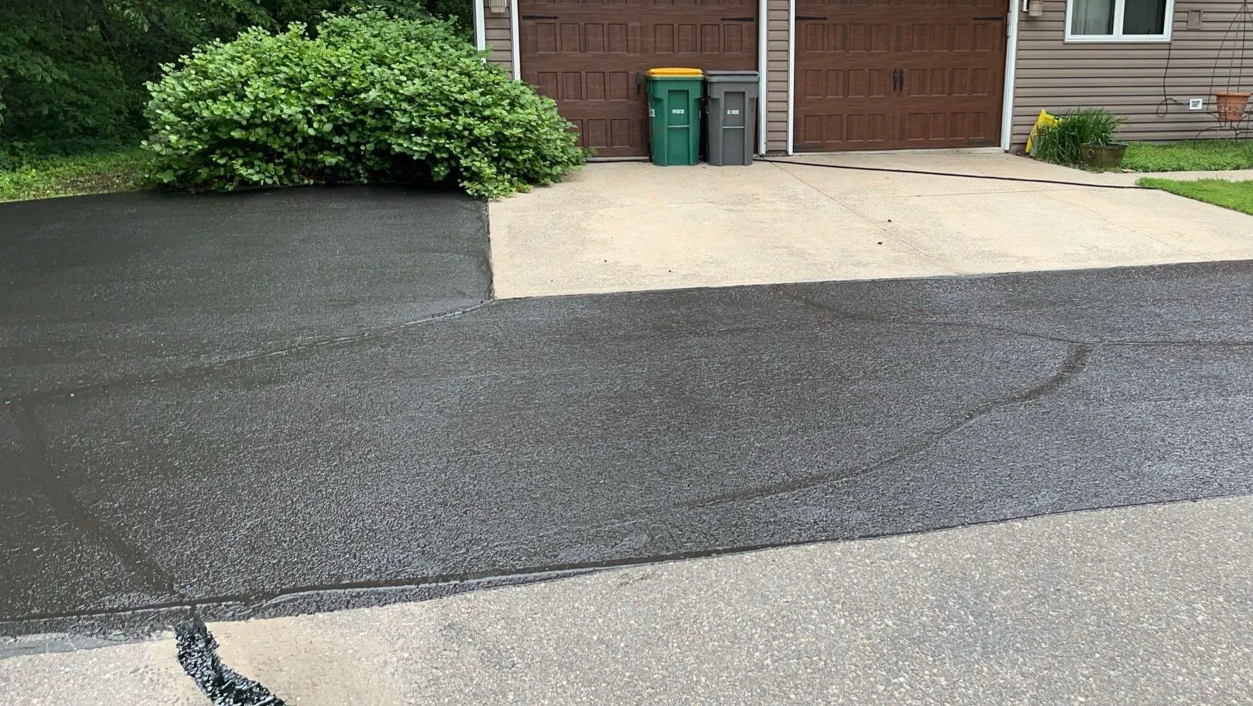 In Process, freshly sealed black asphalt driveway with a section of pressure washed concrete in front of a house.