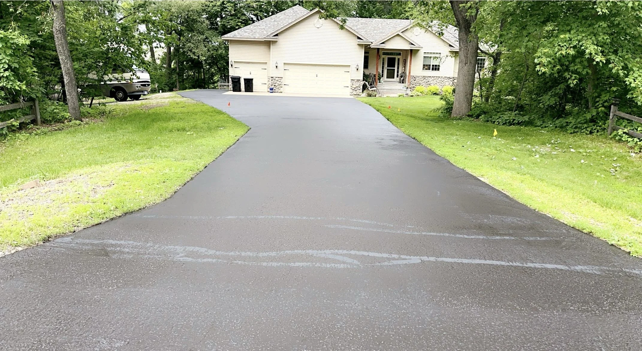 Newly sealed black asphalt driveway leading to a beige house with stone accents, surrounded by green grass and trees, with a black vehicle in the background.