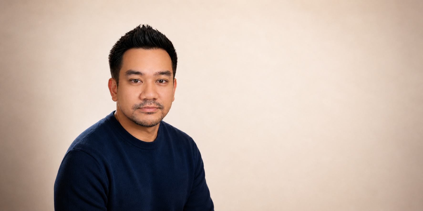 Portrait of a man with short black hair, light facial hair, wearing a navy blue sweater against a plain, light-colored background.