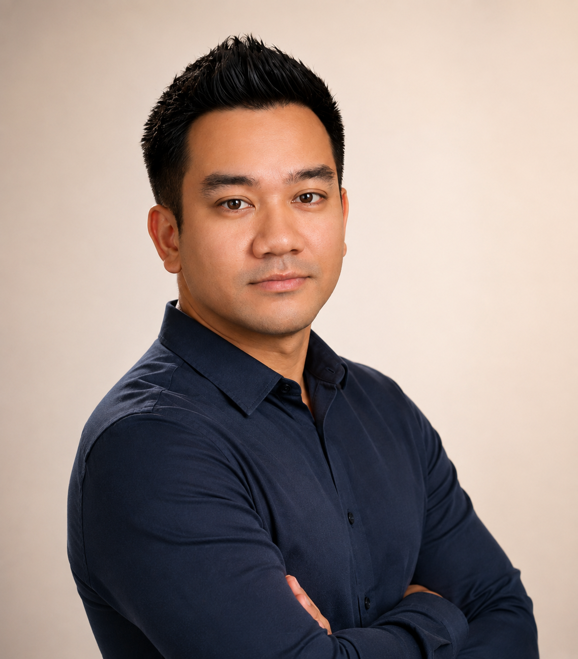 A man with short black hair, wearing a dark navy button-up shirt, posing with his arms crossed against a plain light background.