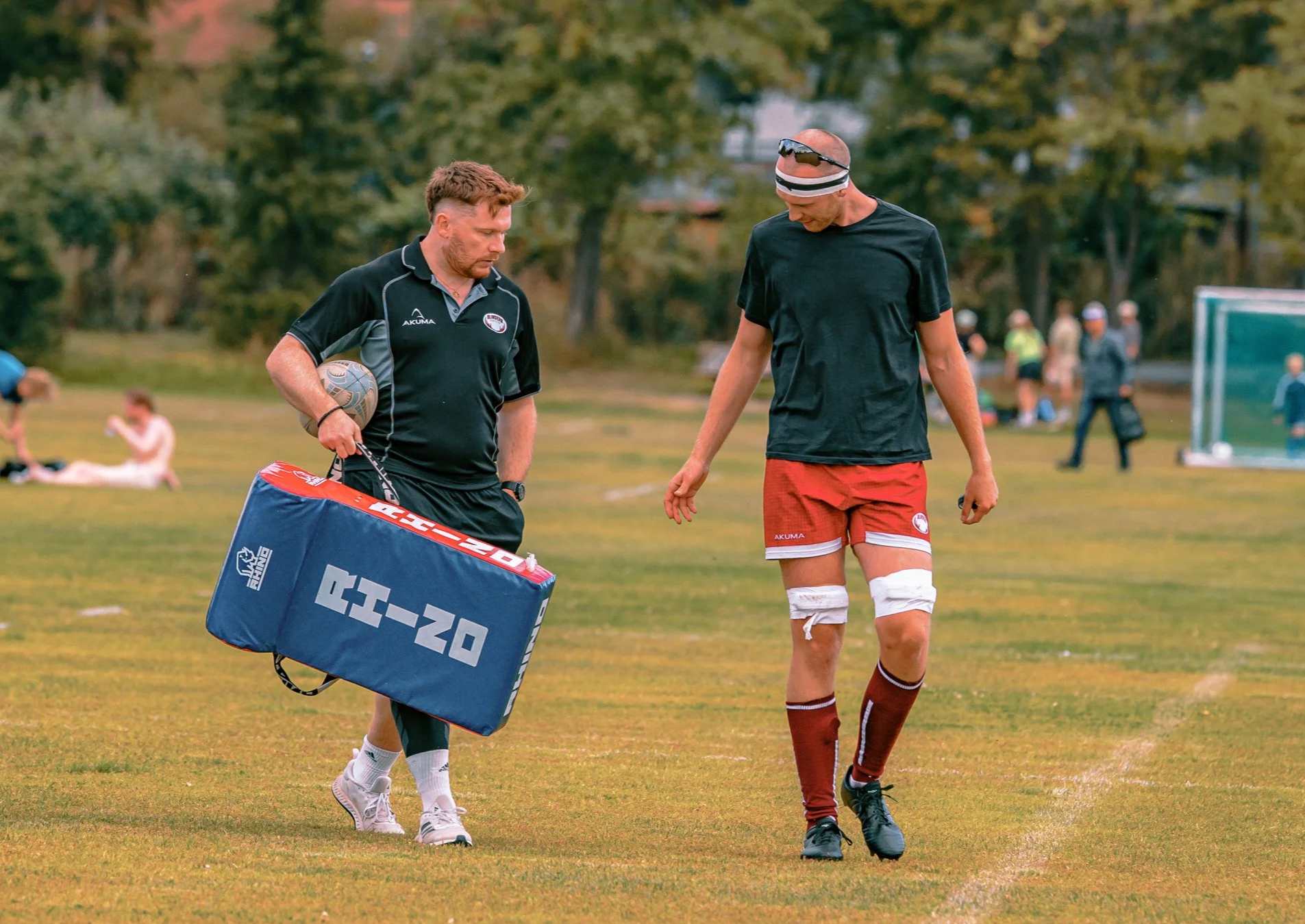 A rugby player in red shorts and black shirt with sunglasses on his head standing on a grassy field, talking to a coach holding a tackle shield and a rugby ball, with other people in the background.