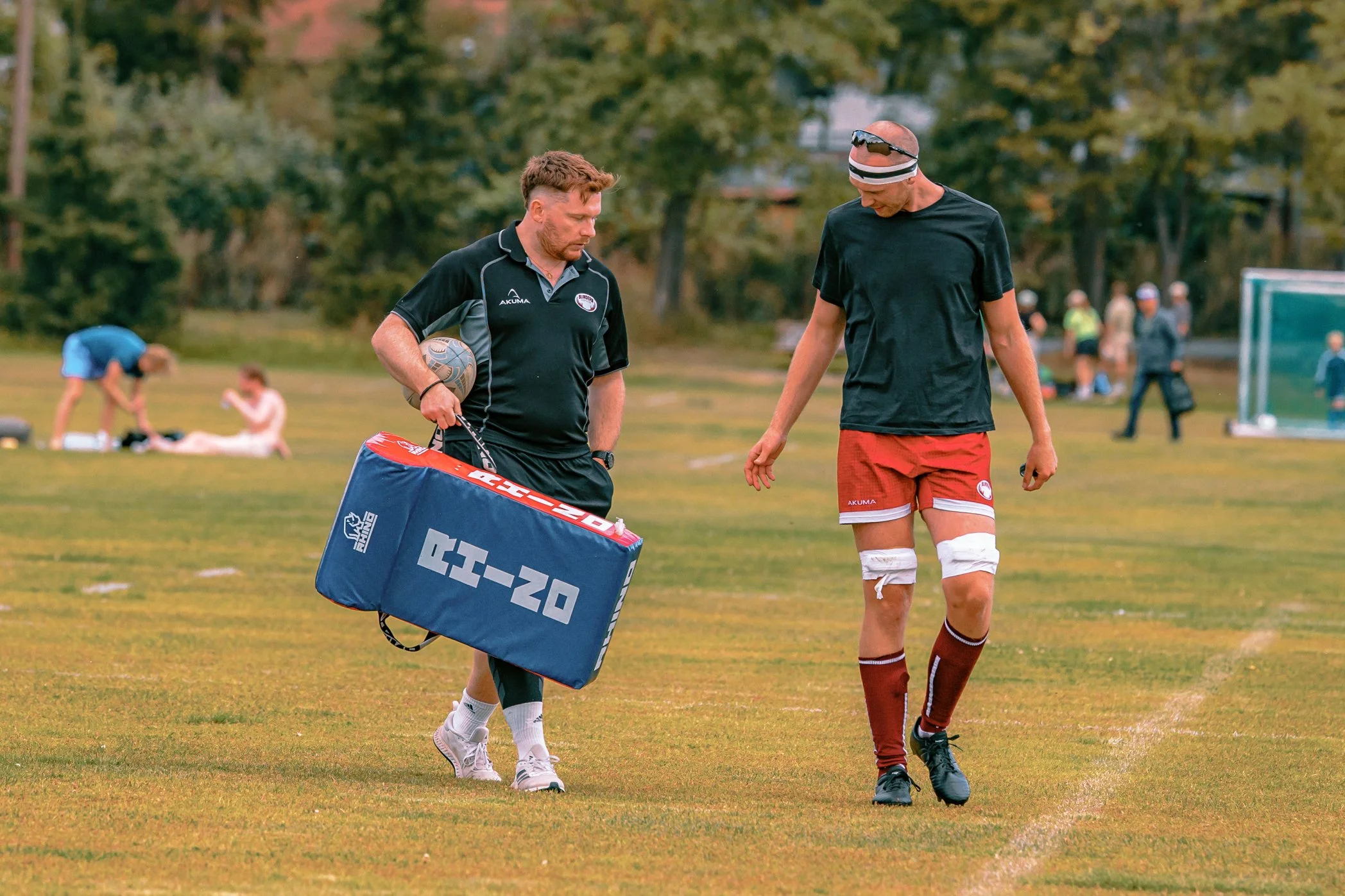 Two men walking on a grass field, one holding a sports bag and the other with a bandage on his knee, with people in the background, possibly after a sports event.