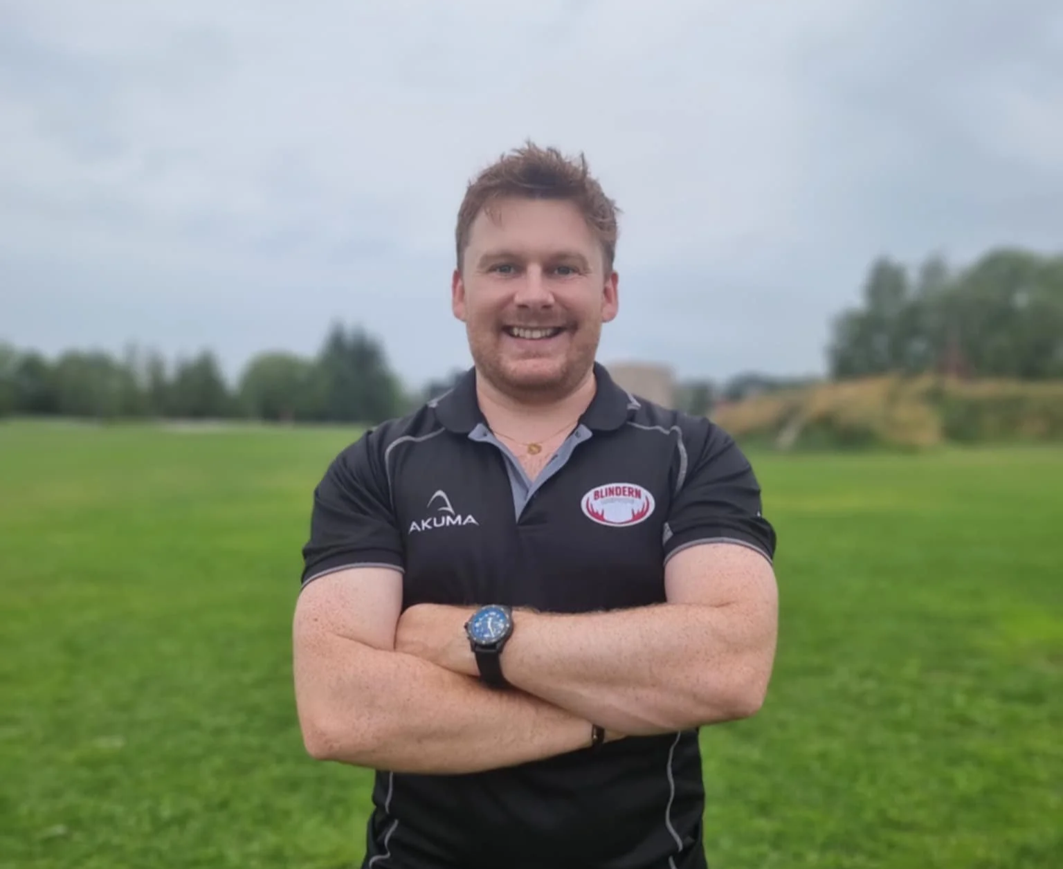 A smiling man with short brown hair, wearing a black sports shirt with a logo that reads 'BLINDERN' and 'AKUMA', standing outdoors on a grassy field with trees and cloudy sky in the background.
