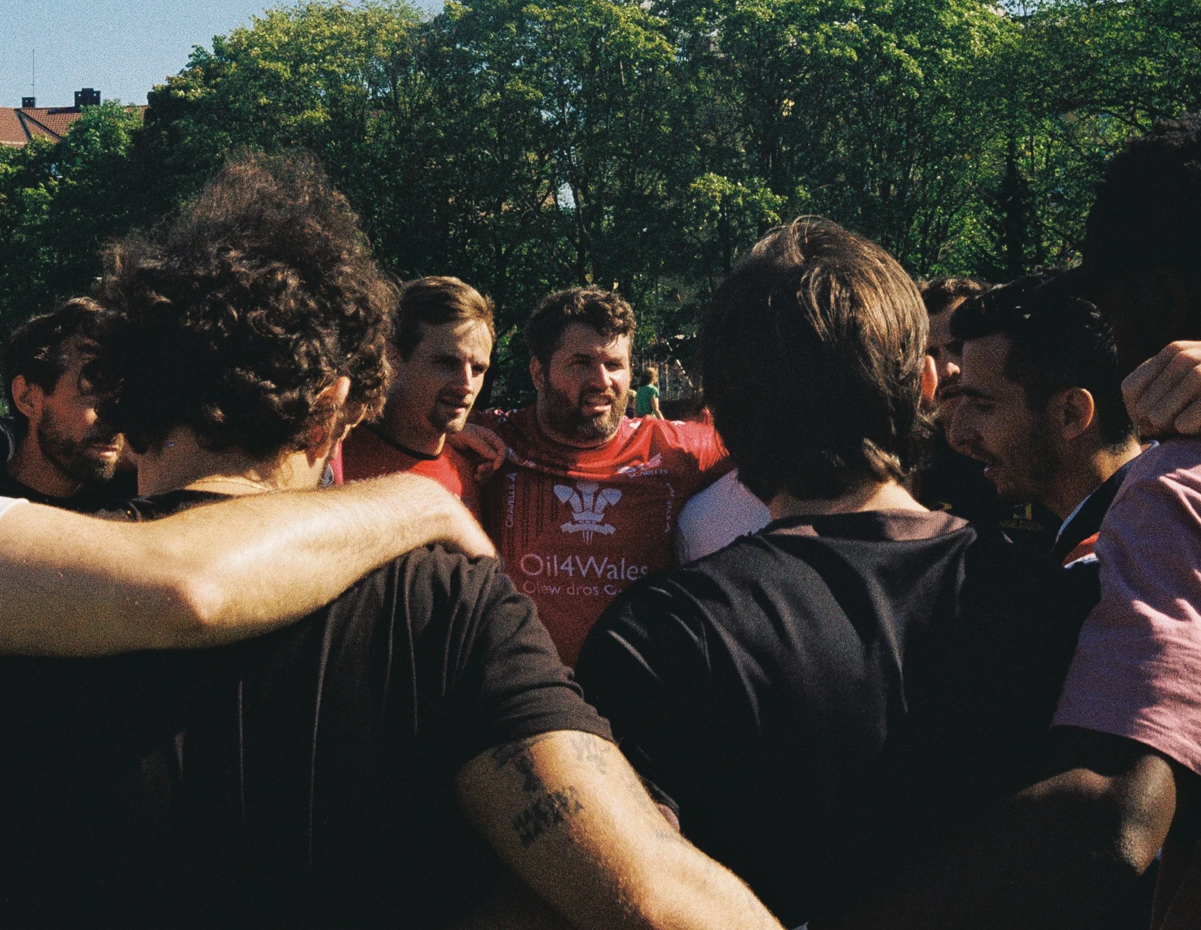 A group of people with their arms around each other forming a huddle outdoors on a sunny day, with trees in the background.