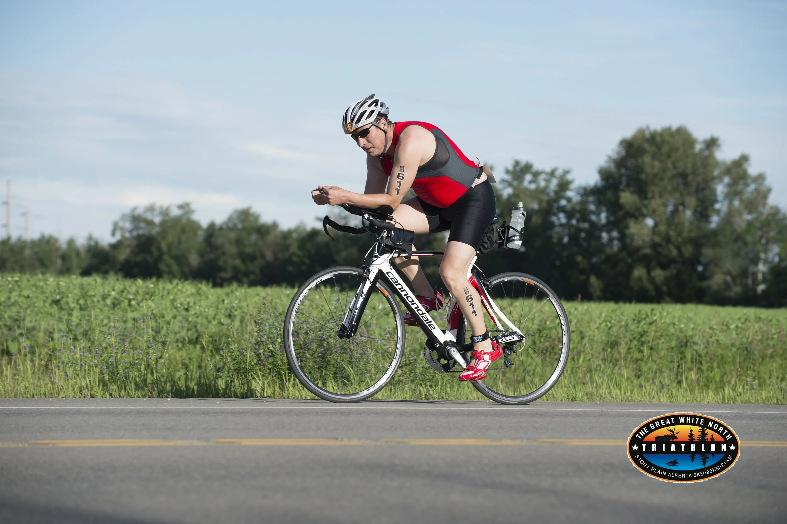 A male cyclist wearing a black and red sleeveless shirt, black shorts, red cycling shoes, and a helmet, riding a Cannondale road bike on a paved road with green fields and trees in the background. There is a logo in the bottom right corner that says "The Great White North Triathlon" with additional details.