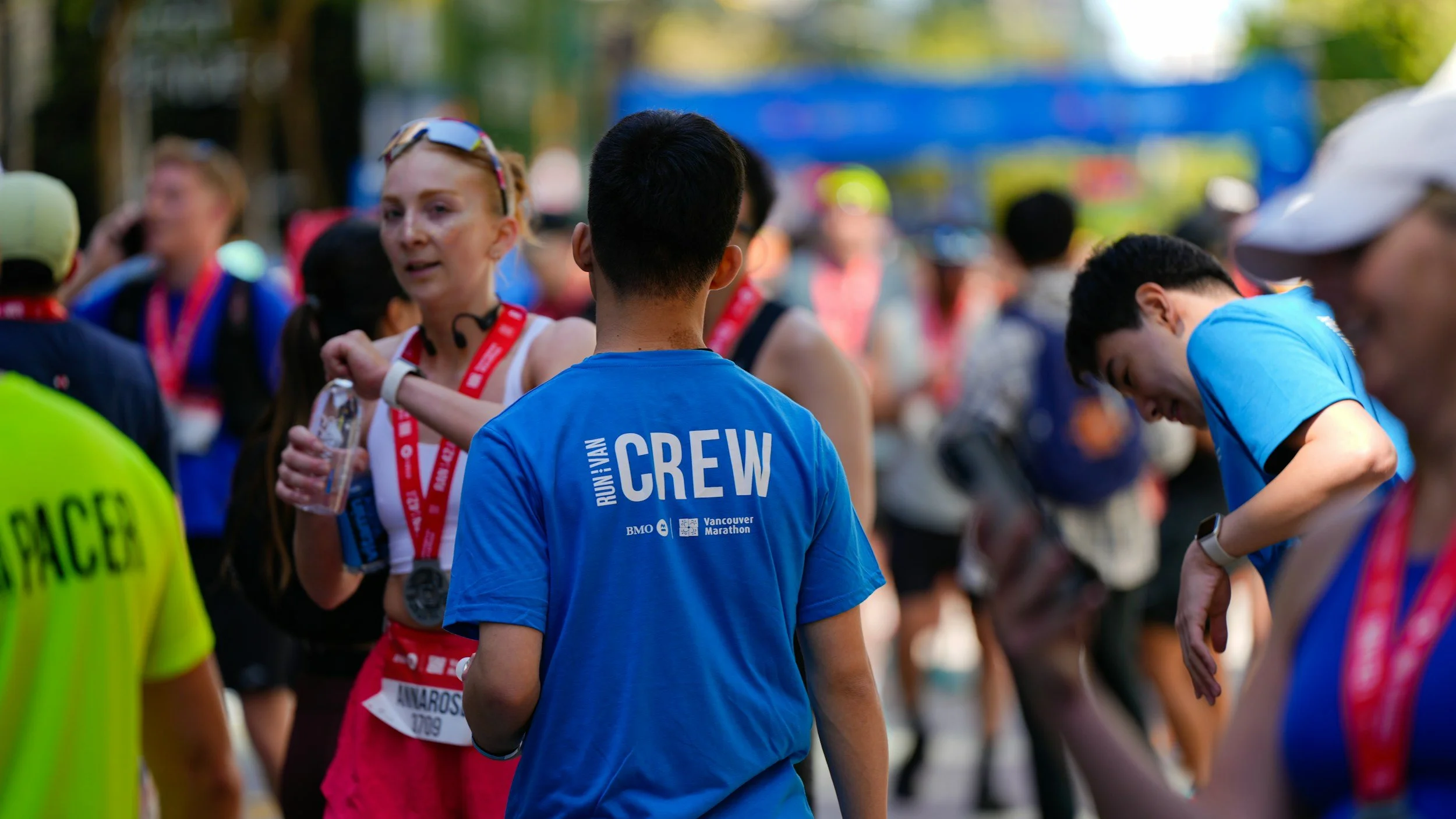 Group of marathon runners gathering before the race with volunteers and race staff around, some wearing medals and race gear, in a tree-lined park.