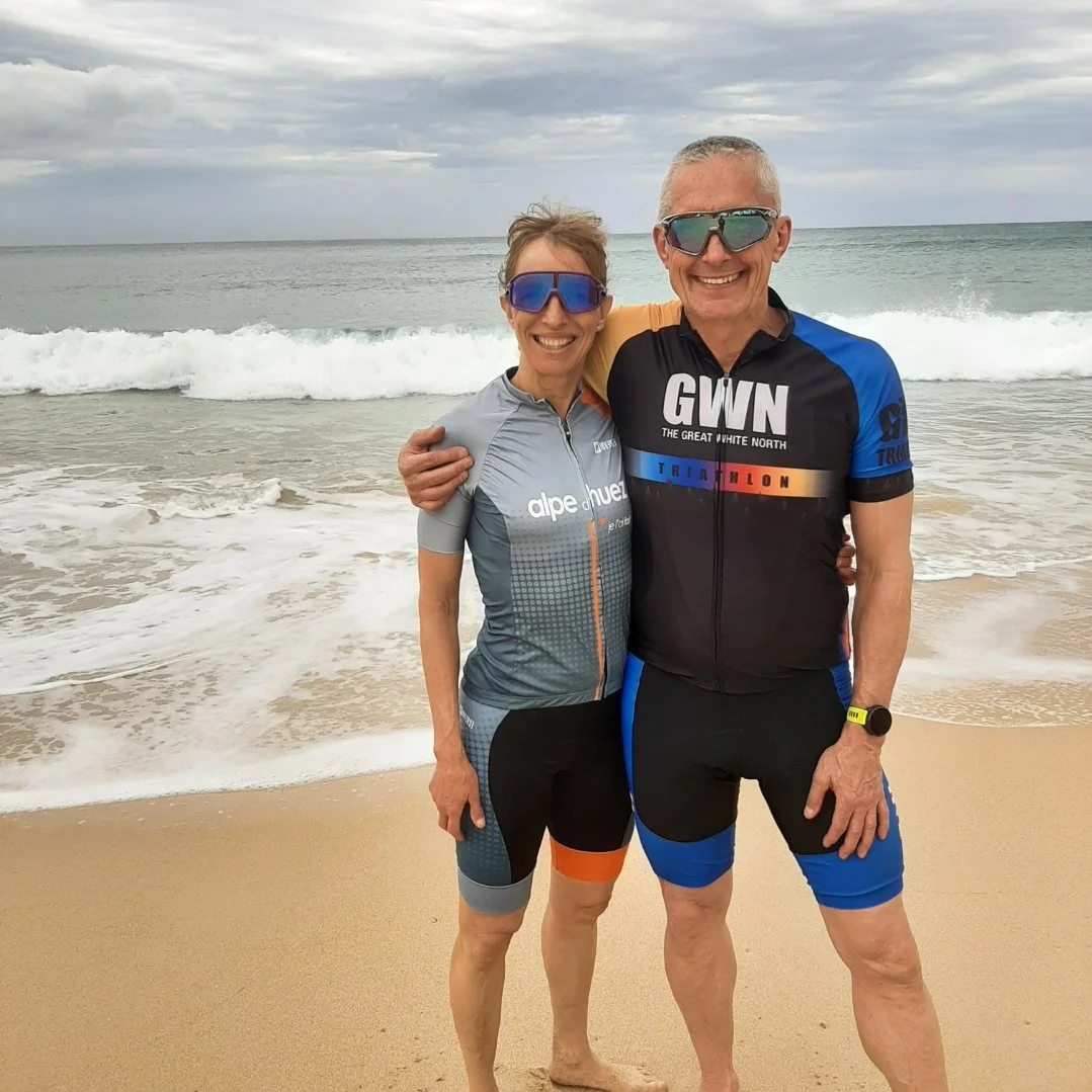 A smiling middle-aged man and woman wearing cycling gear and sunglasses, standing on the sandy beach with ocean waves behind them.