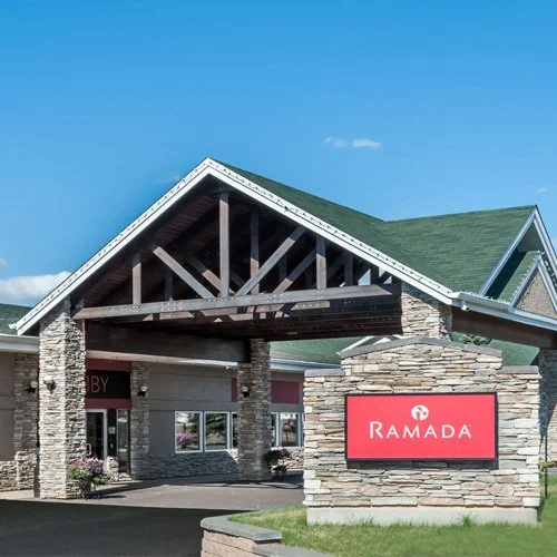 Ramada hotel entrance with a stone and wood facade, green roof, and a large red Ramada sign in front.