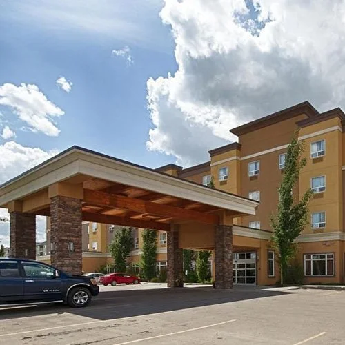 Exterior view of a modern hotel or apartment building with a large covered entrance supported by stone pillars, parking lot with cars, and a partly cloudy sky.