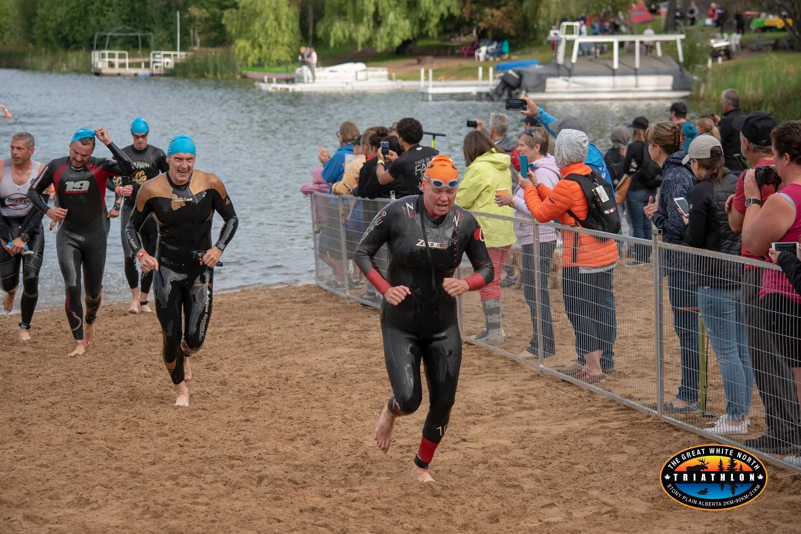A group of triathletes exiting the water and running onto the beach during a race, with spectators taking photos and cheering along the shoreline, near a lake with boats and trees in the background.