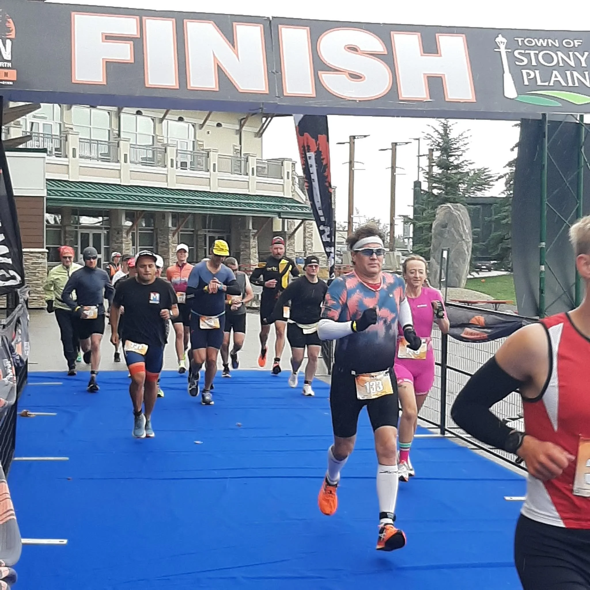 Group of marathon runners crossing the finish line outdoors, with a large sign overhead that reads 'FINISH' and a picture of a lamp post and the words 'Town of Stony Plain'.