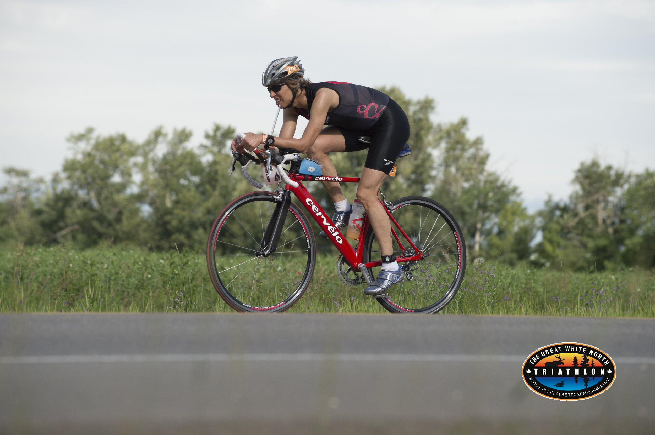Female triathlon athlete in black and red outfit riding a red Cervélo road bike on a grassy area during a race, with a forested background and a triathlon logo at the bottom right.