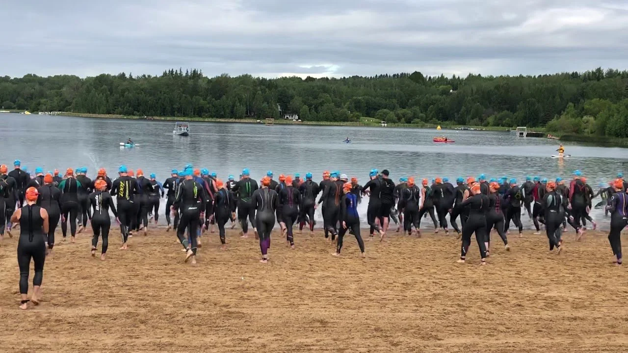 Many triathletes in wetsuits and orange and blue swim caps entering a lake on a sandy beach, with trees and cloudy sky in the background.